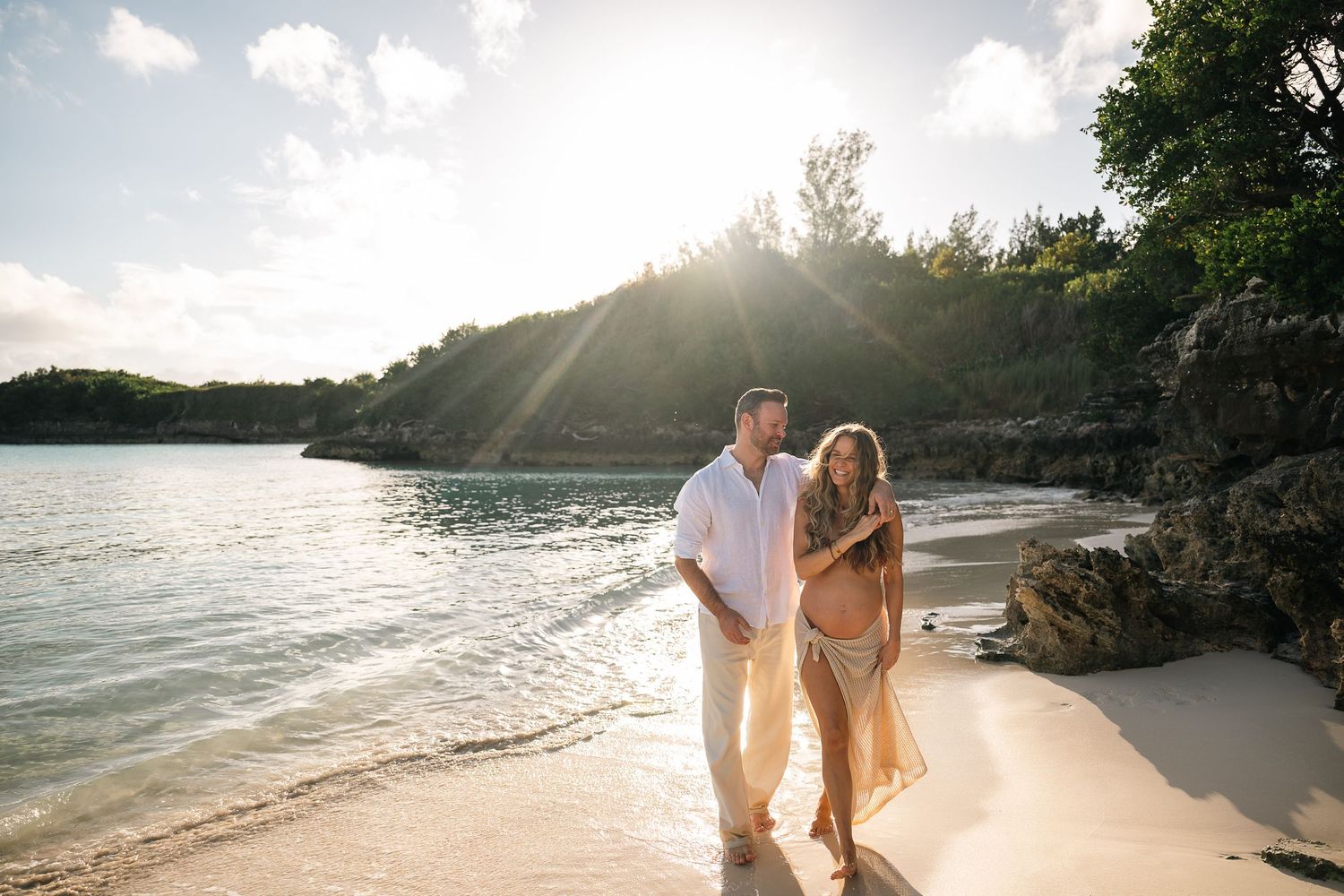 Happy couple walking hand in hand along a sunny tropical beach at golden hour with lush greenery behind.