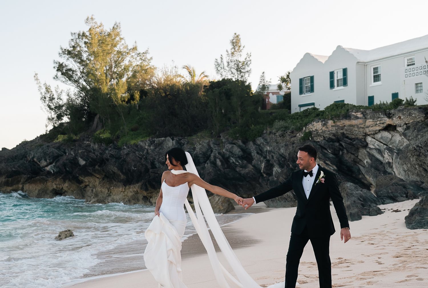 Bride and groom holding hands on beach at dusk, veil flowing in wind with rocky cliffs and white house behind.