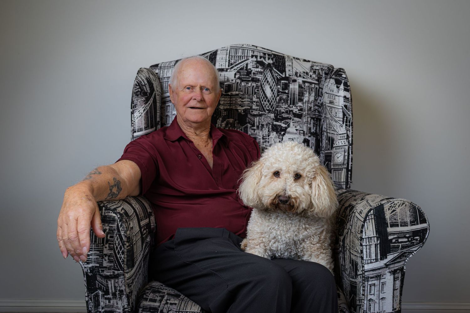 Older man seated in a patterned armchair with his white curly coated dog