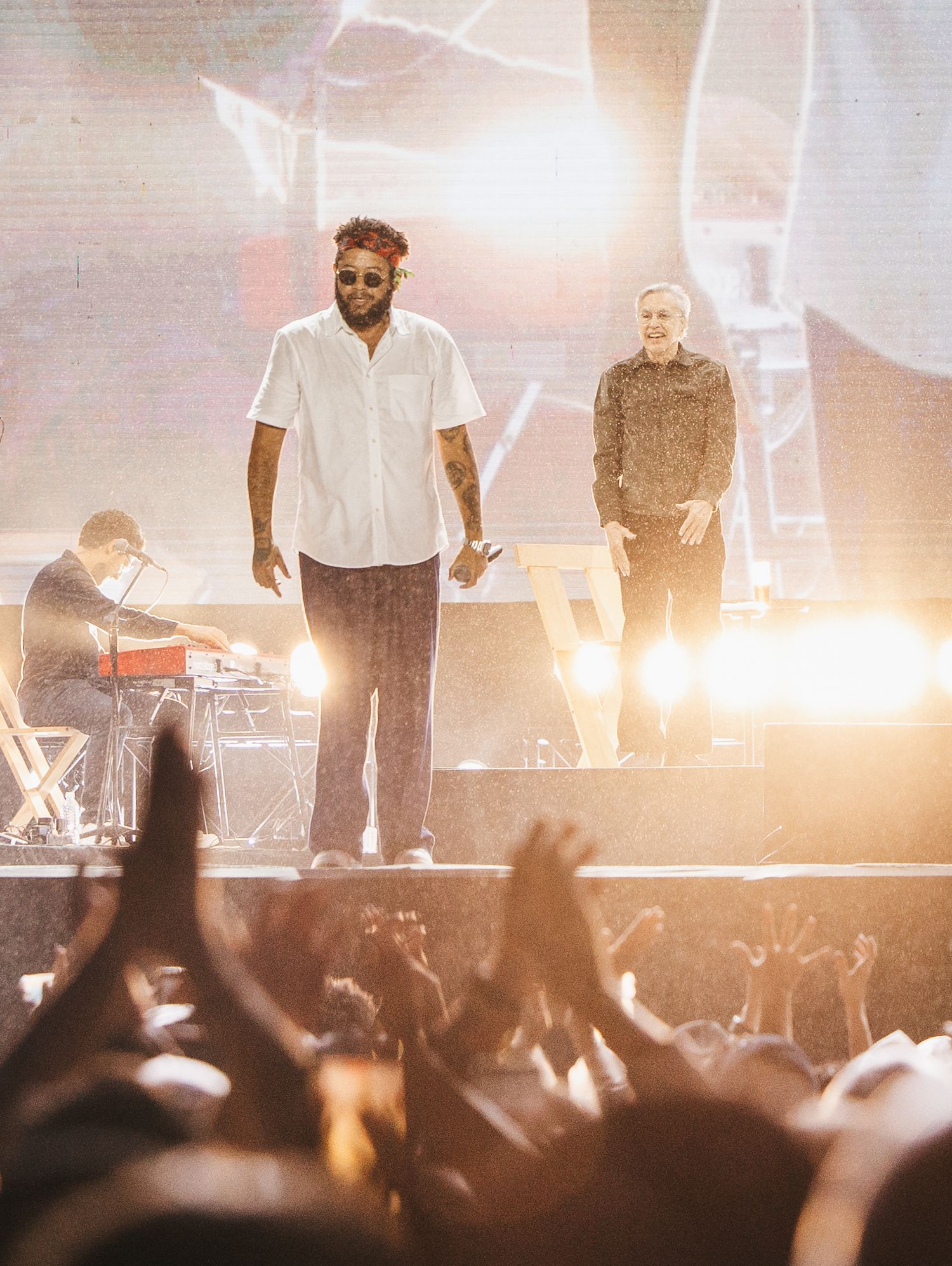 Two performers on a bright concert stage engage an enthusiastic crowd with raised hands, while a keyboardist plays in the background.