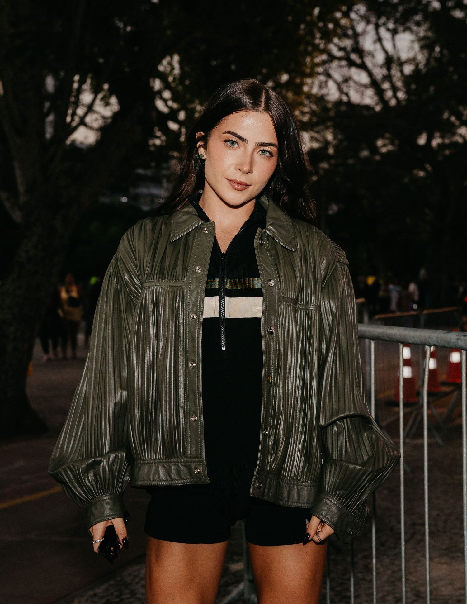 Young woman in olive green leather jacket and black outfit posing outdoors at night near metal barriers.