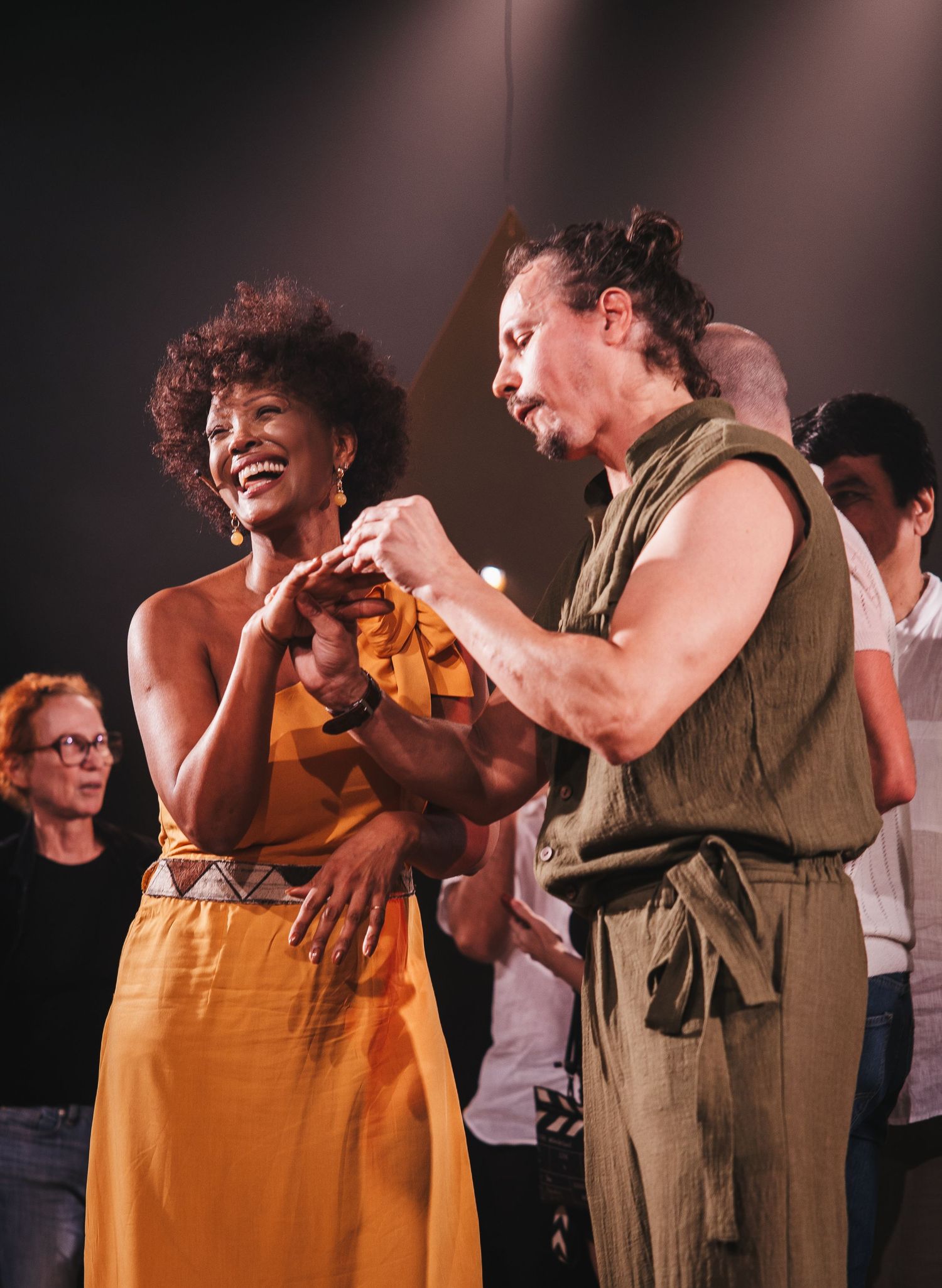 A woman in a yellow dress and a man in olive green dance joyfully together on a theater stage.