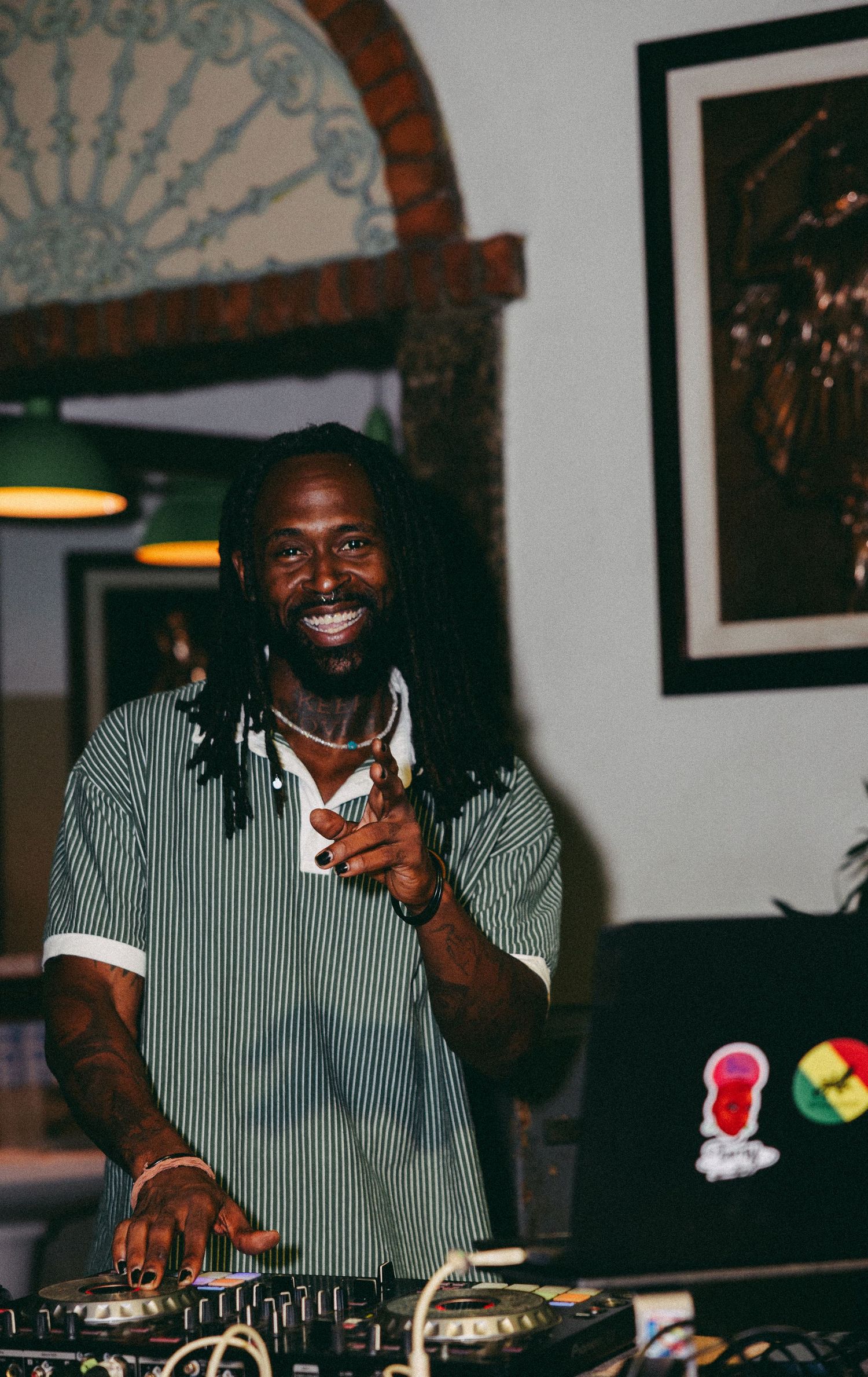 A smiling DJ with dreadlocks works the turntables at an indoor music event, pointing at the camera.