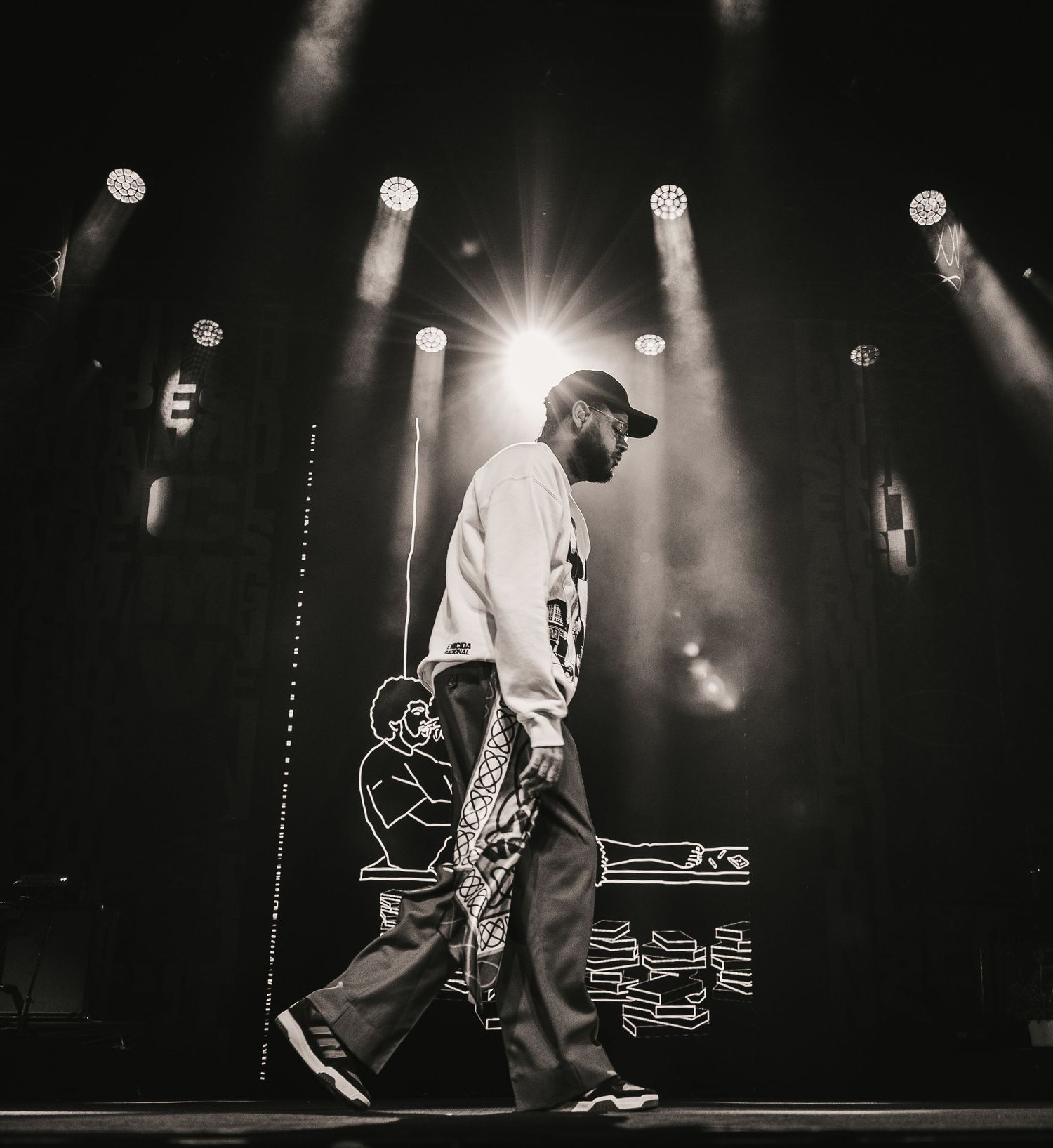 Black and white silhouette of a performer walking across a stage with bright spotlights behind.