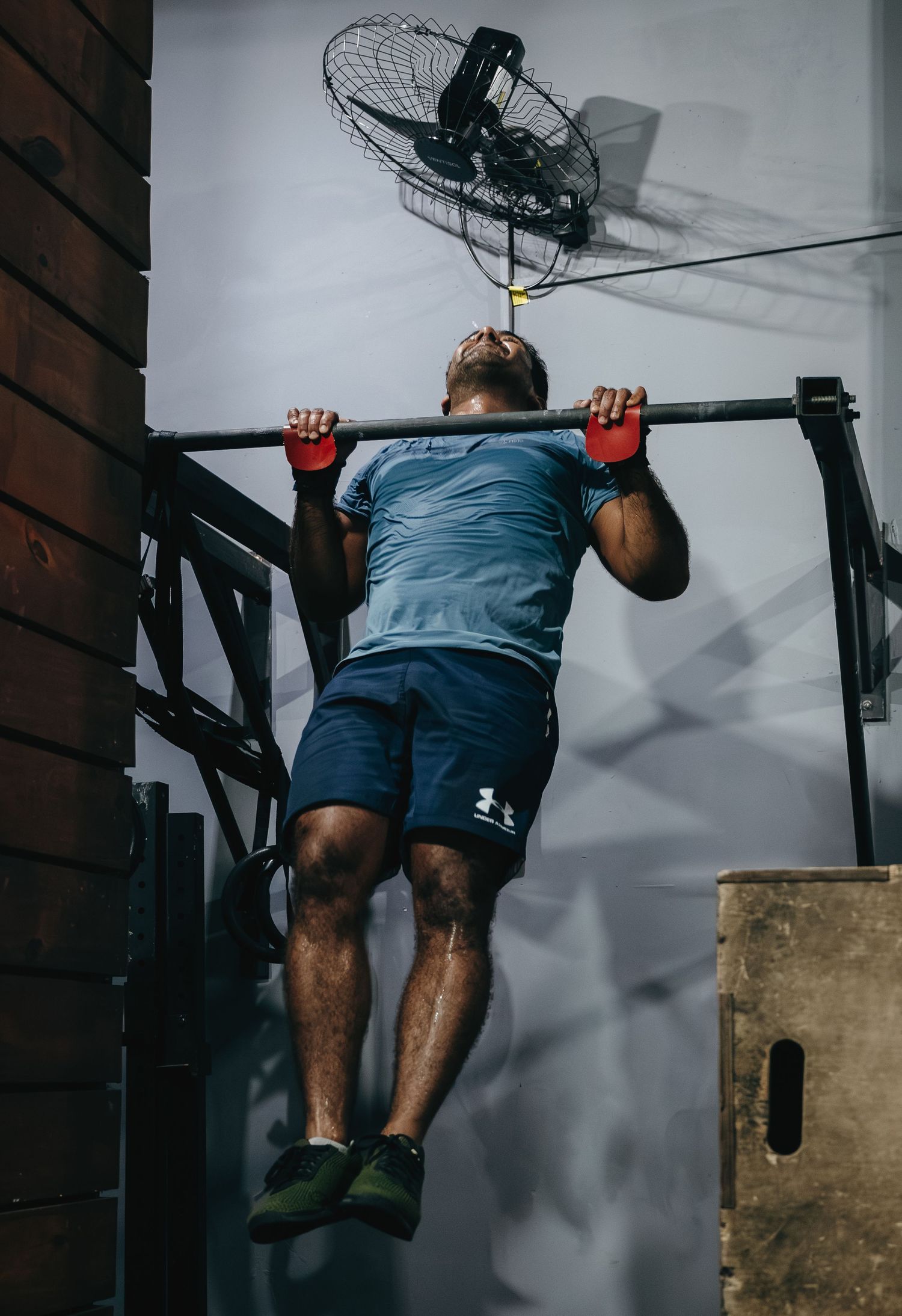 Man doing pull-ups on a bar in a gym wearing Under Armour shorts and red gloves with a wall fan above.