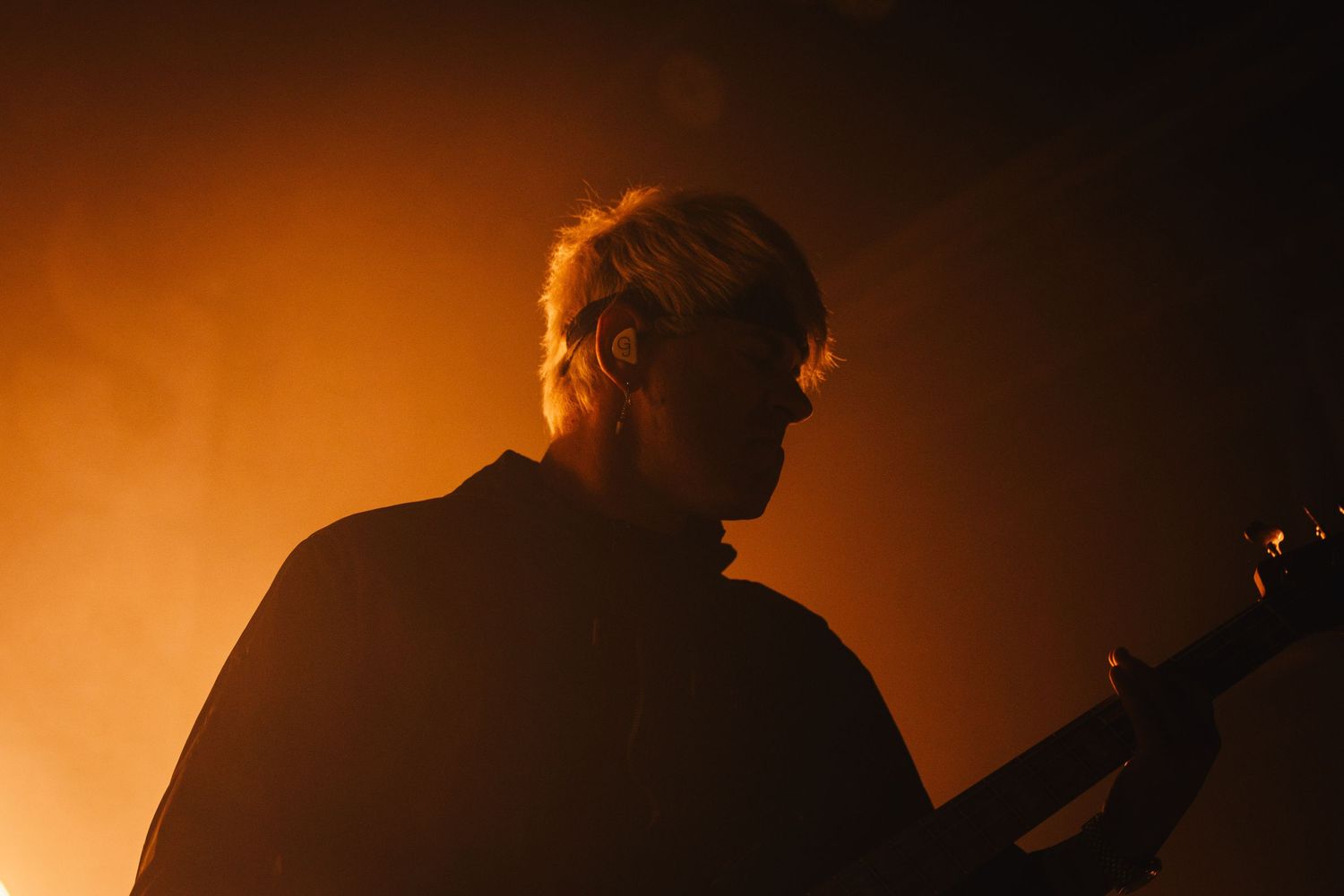 Silhouetted guitarist performing on stage bathed in warm orange atmospheric fog lighting.