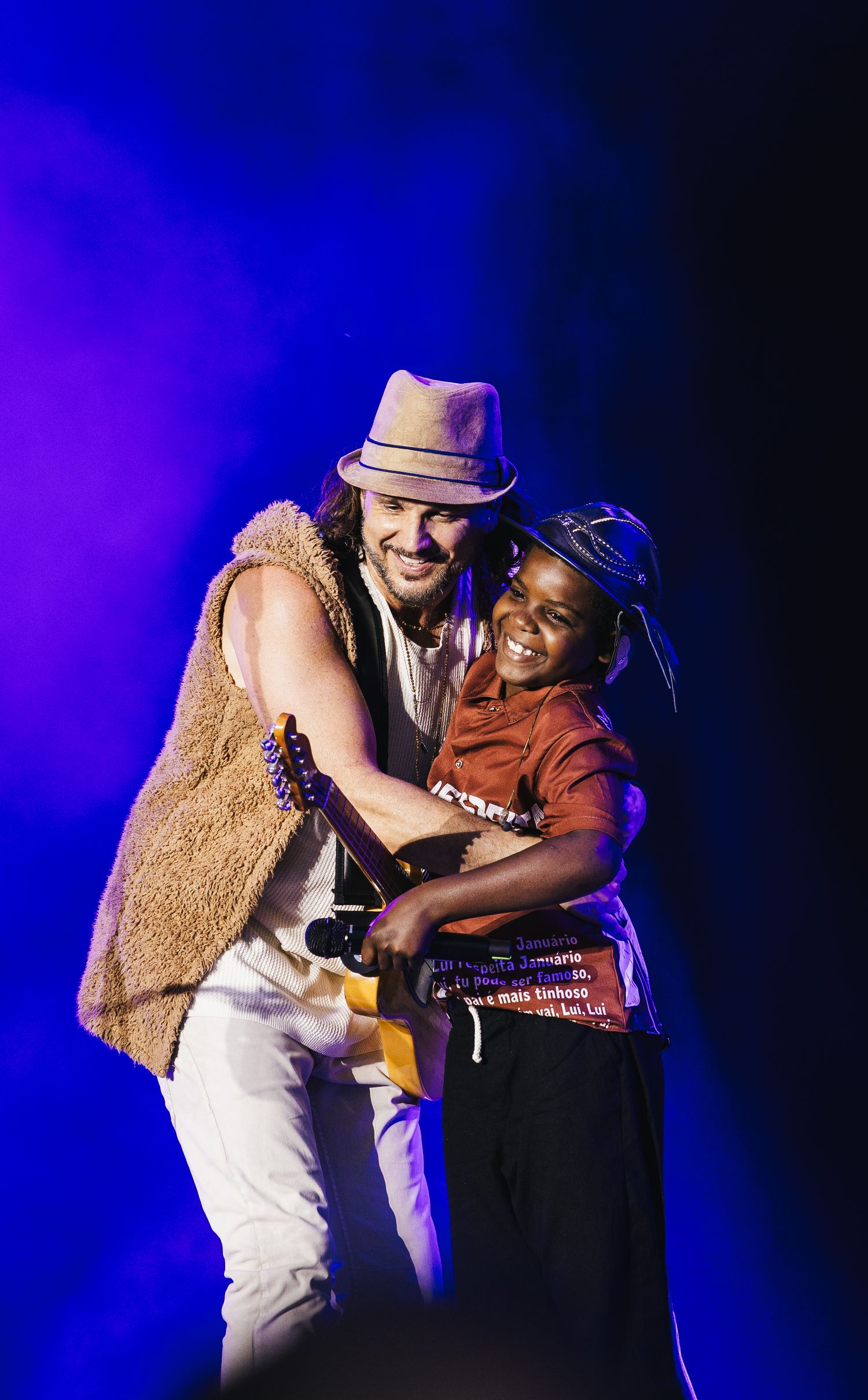 Musician in hat hugs smiling child on stage during live concert performance with blue stage lighting.