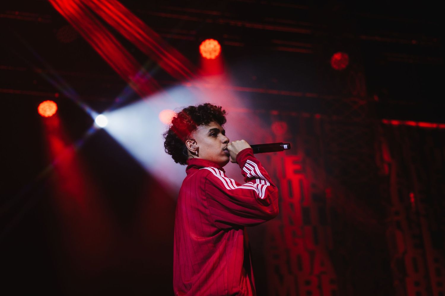 Young male singer in red tracksuit performs on stage under red spotlights at a live show.