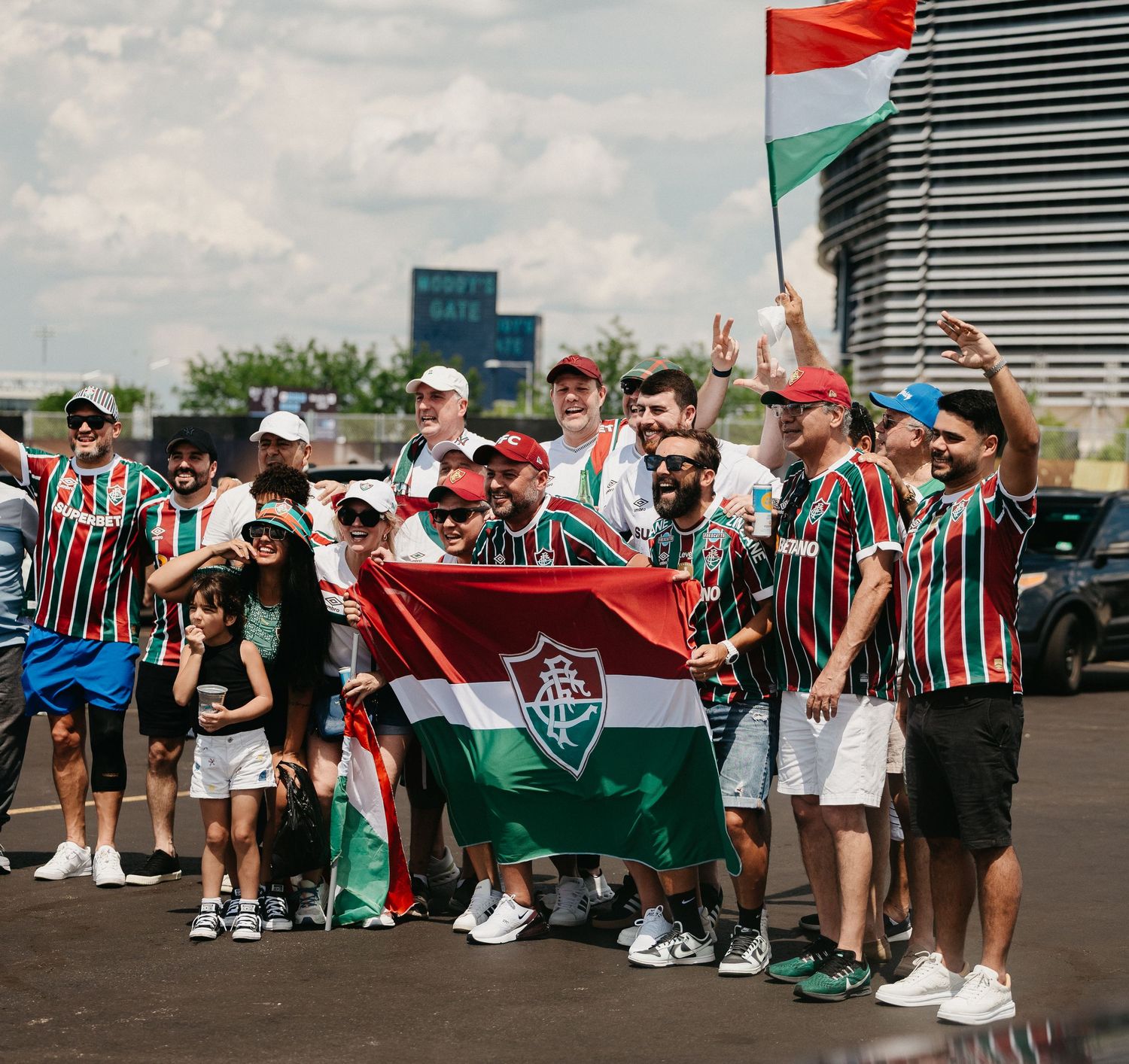 Group of Fluminense soccer fans holding flags and celebrating outdoors on sunny day.