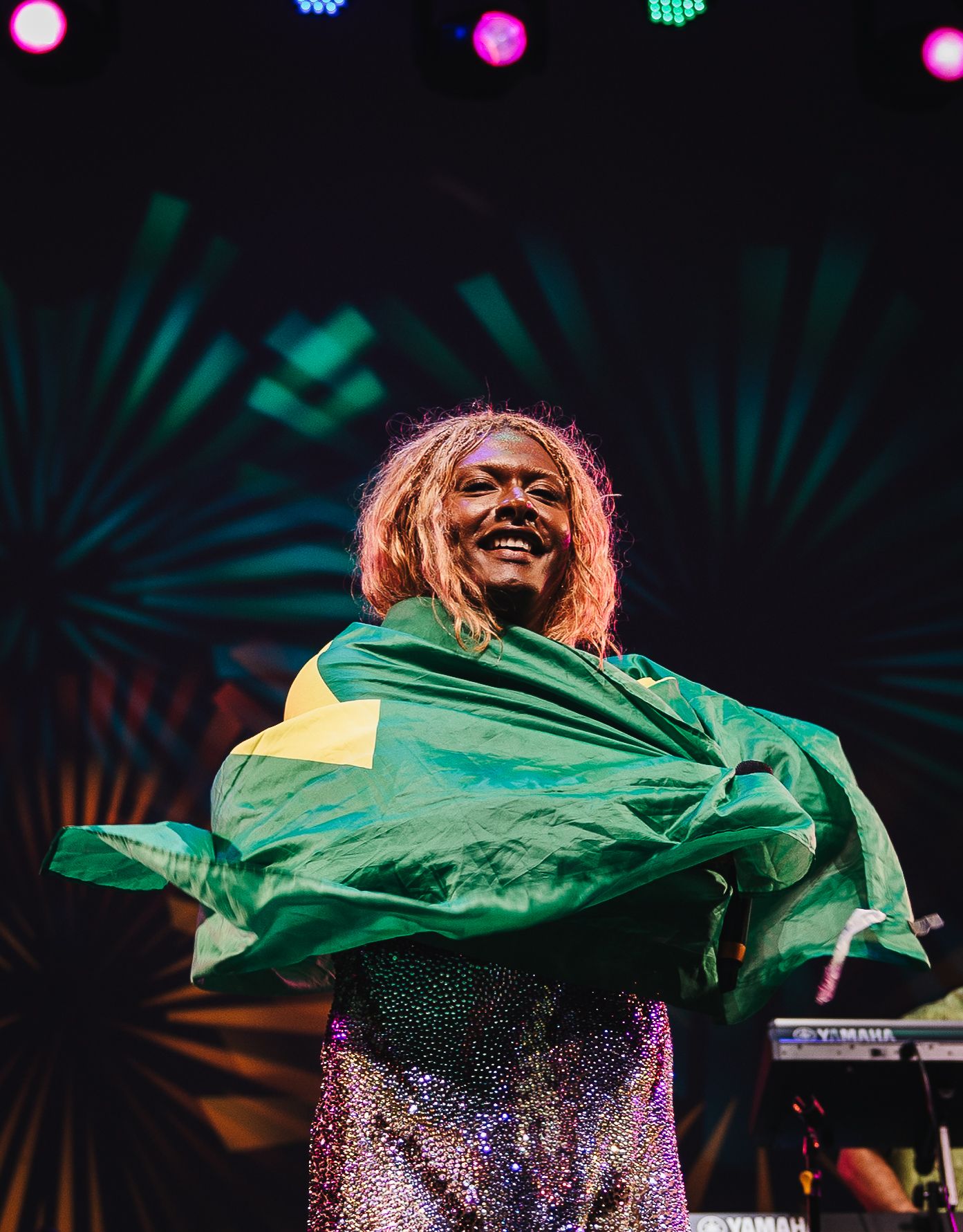 Smiling performer on stage wrapped in Brazilian flag, wearing sequined outfit with colorful lights in background.