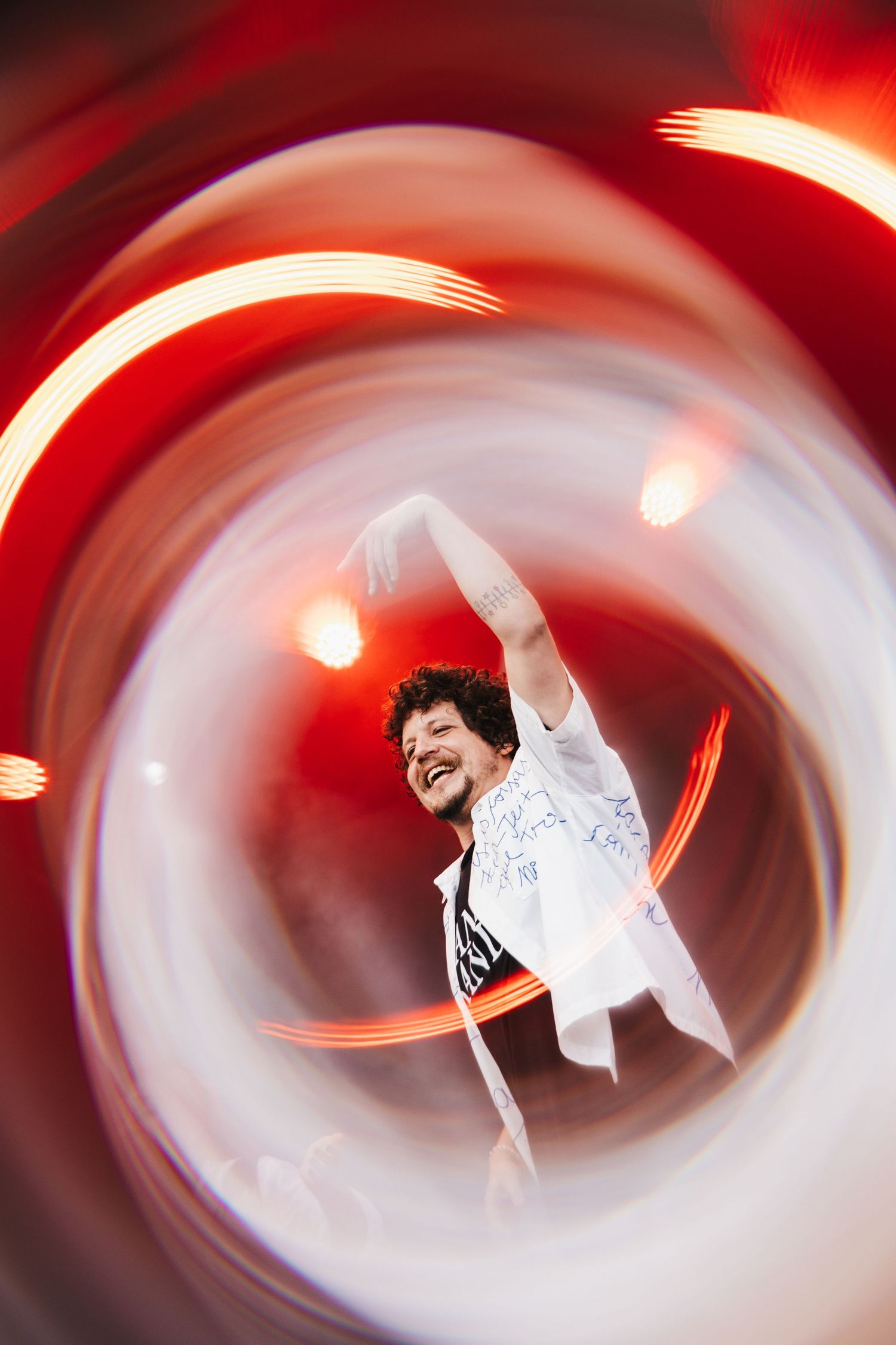 A man in a white shirt dances joyfully, surrounded by swirling red and white light trails at a concert.