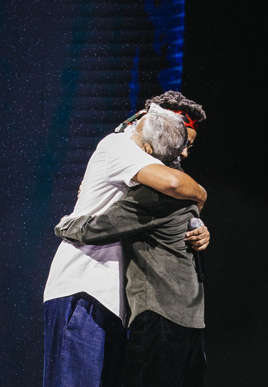 Two people sharing a warm embrace on a dimly lit stage during a live performance event.