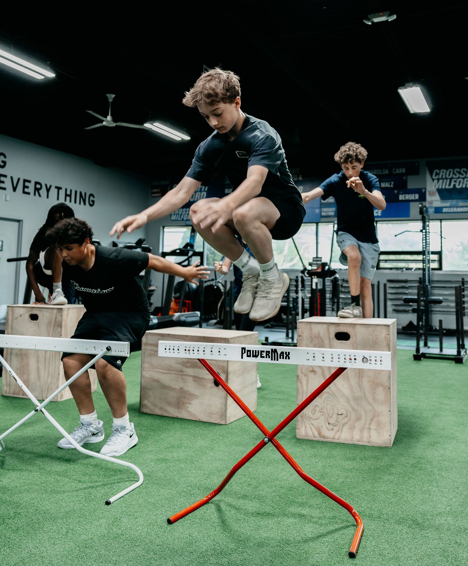 Teen athletes perform box jumps on PowerMax plyo boxes in a modern CrossFit gym during a youth training session.