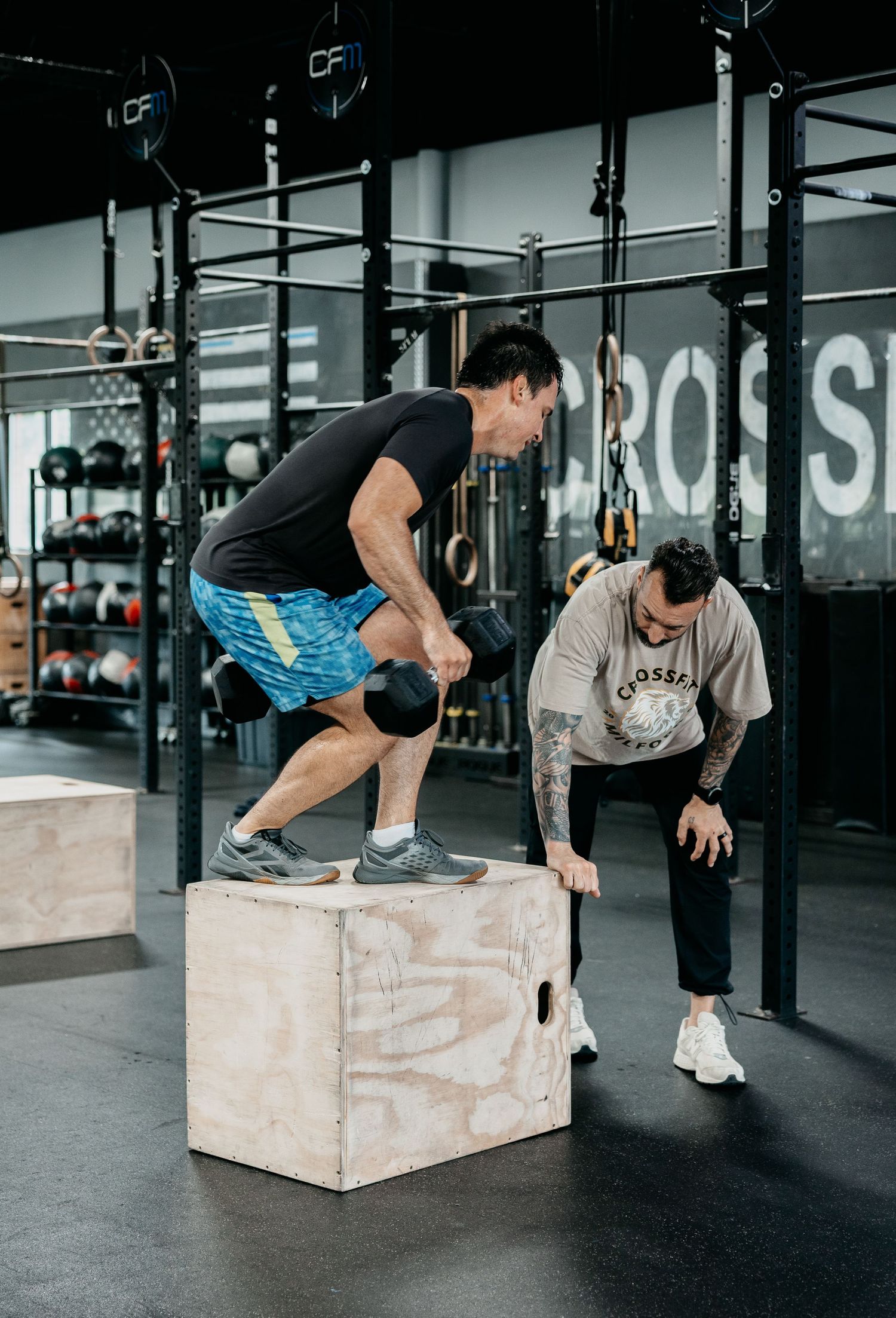 Two men training at a CrossFit gym, one performing a kettlebell box step-up while the other coaches nearby.
