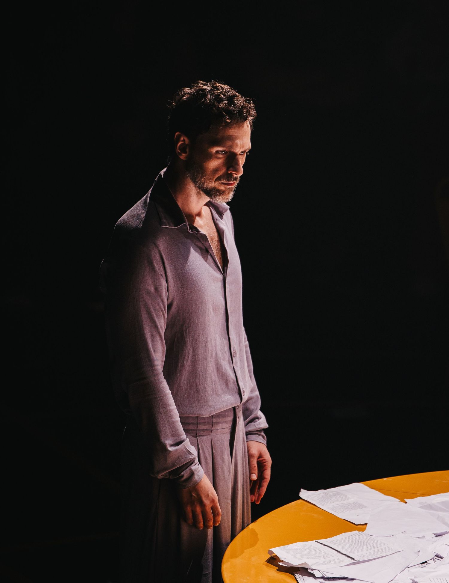 A man in a gray shirt stands pensively over a yellow table with papers in dramatic stage lighting.
