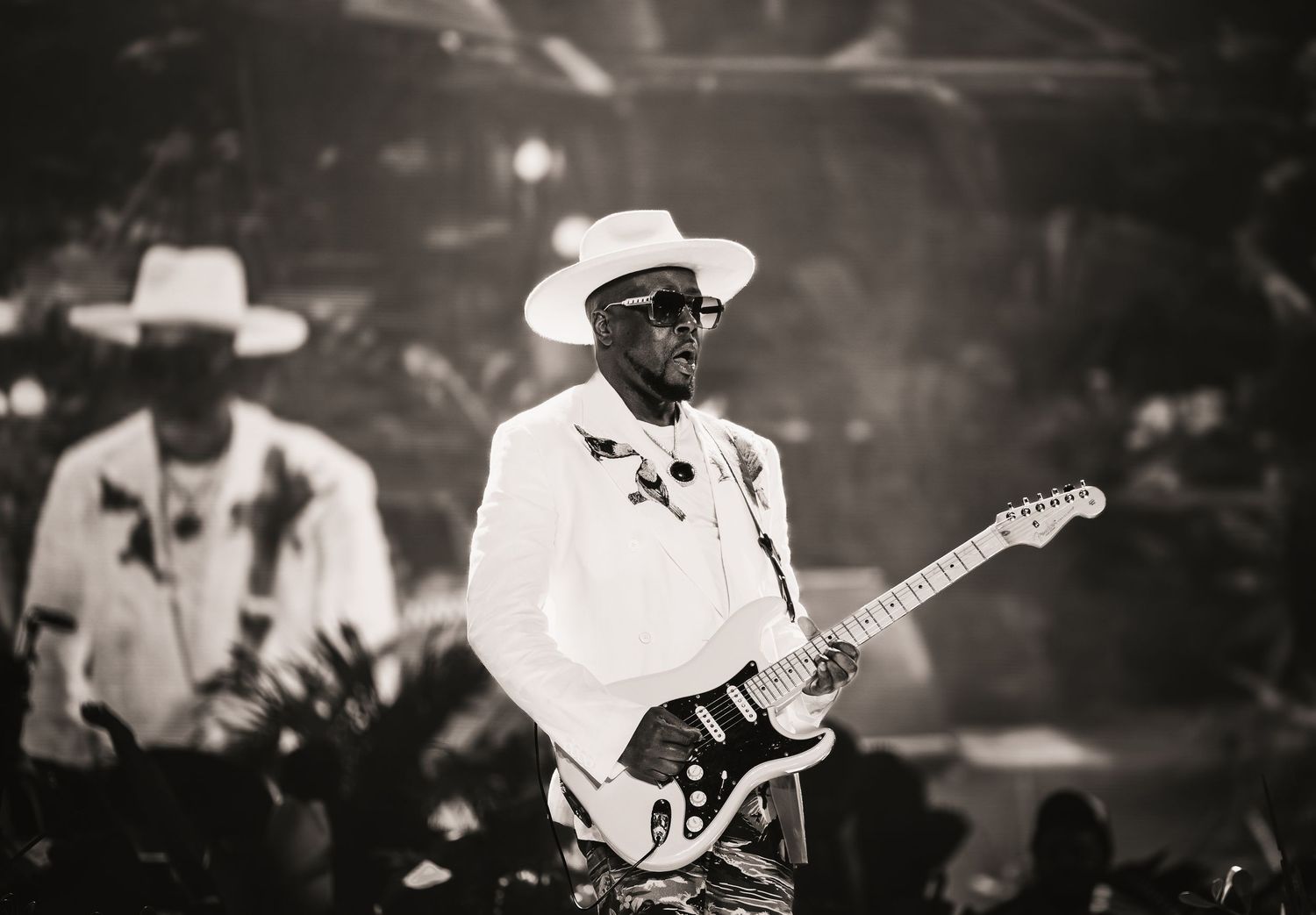 Black and white photo of a guitarist in white suit and hat performing on stage with another musician in background.