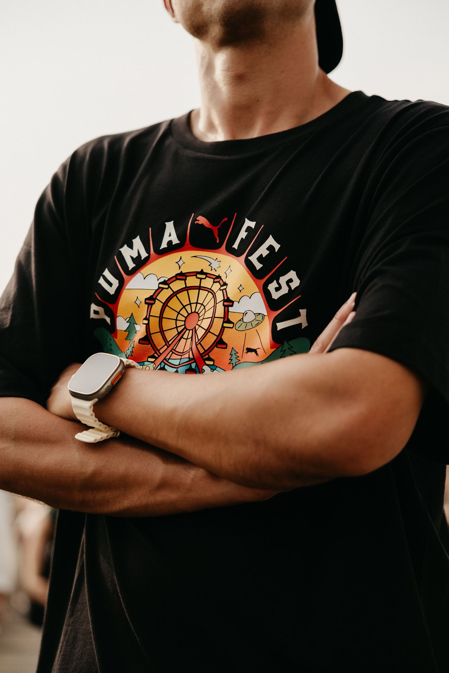 Man with arms crossed wearing a black Puma Fest graphic t-shirt featuring a ferris wheel design.