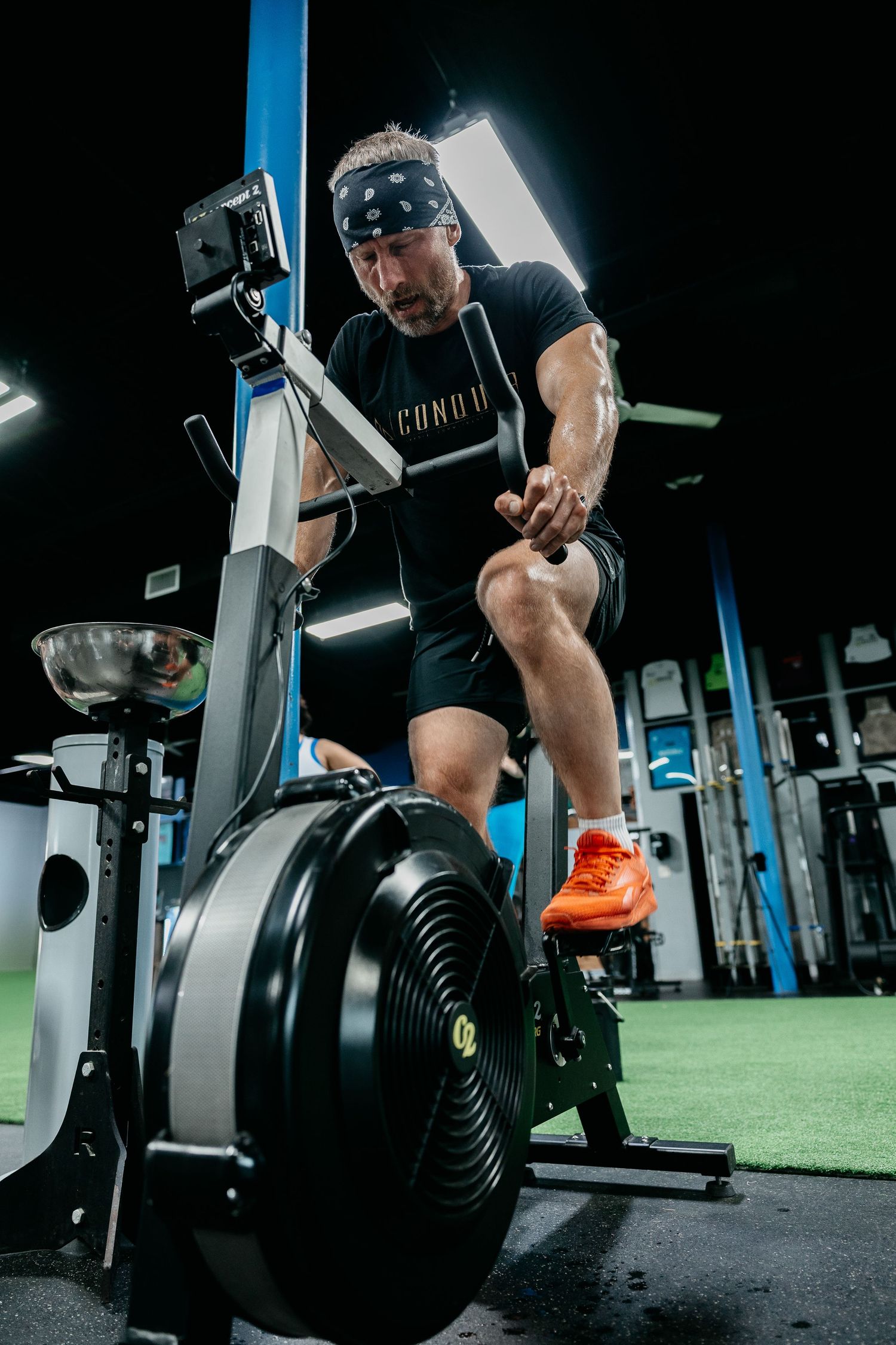 Man in black shirt and bandana intensely working out on an air bike machine in a dark gym setting.
