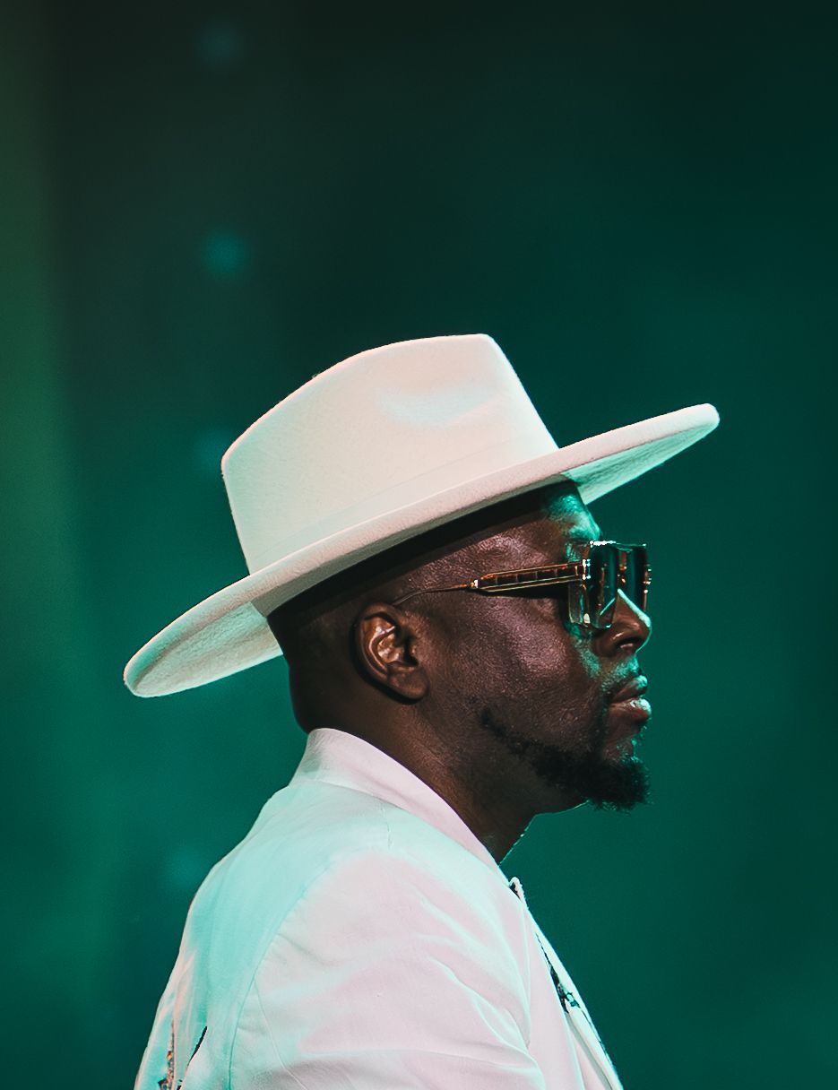 Man in white wide-brim hat and stylish glasses performing on stage against a teal background.