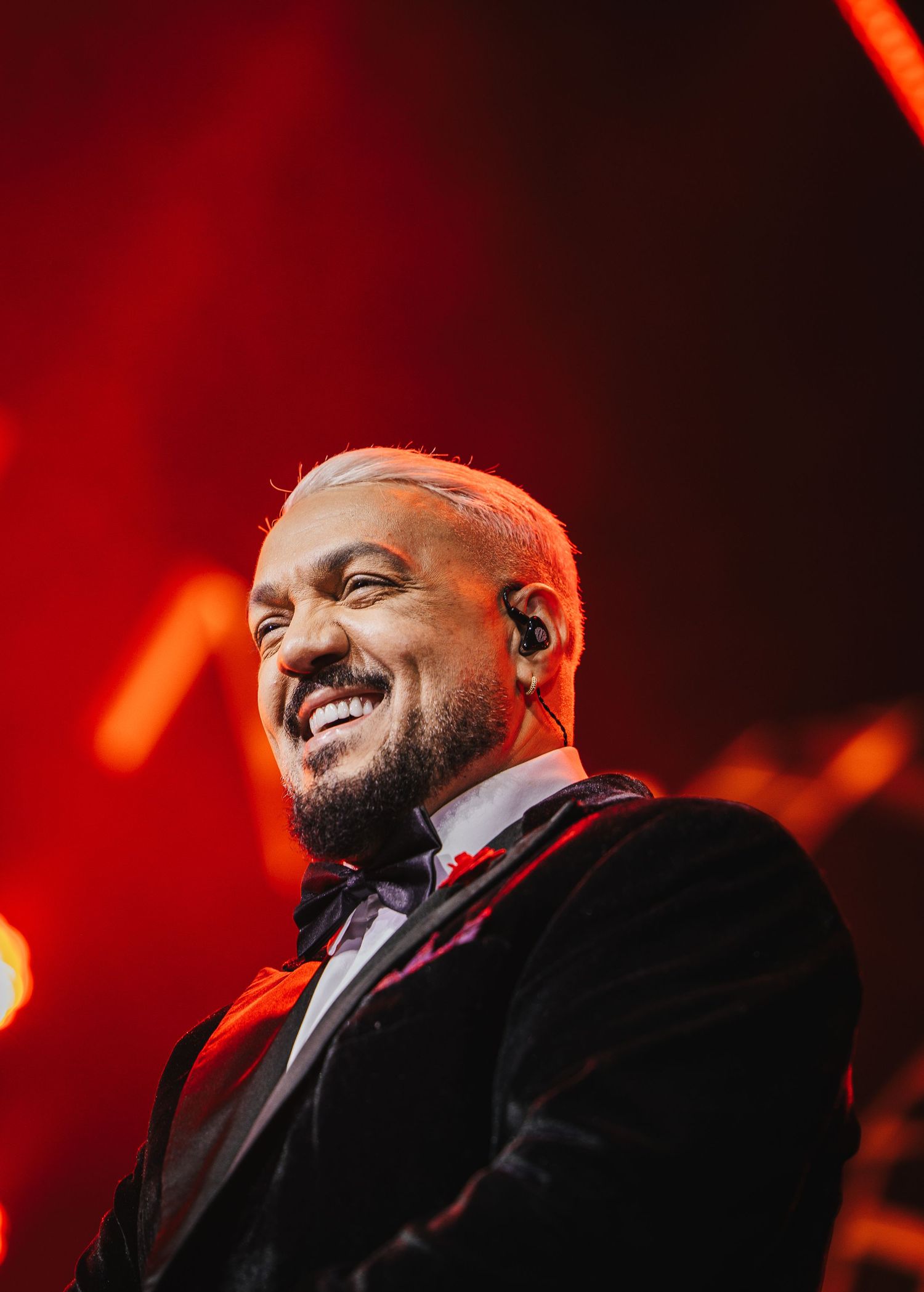 Smiling male performer in black velvet tuxedo with bow tie on stage, bathed in red concert lighting.