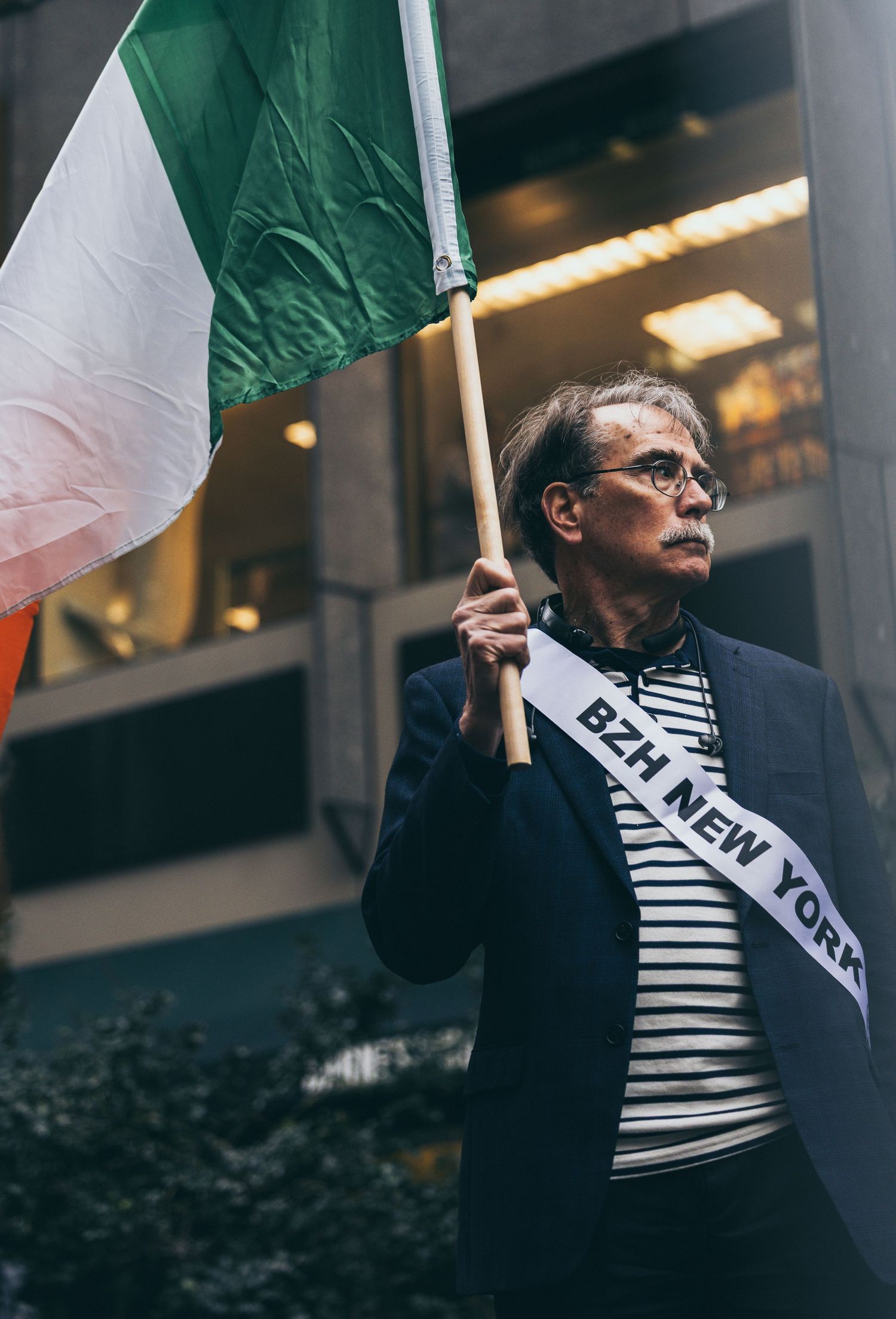 Man holding an Irish tricolor flag wearing a BZH New York sash at an outdoor parade event.