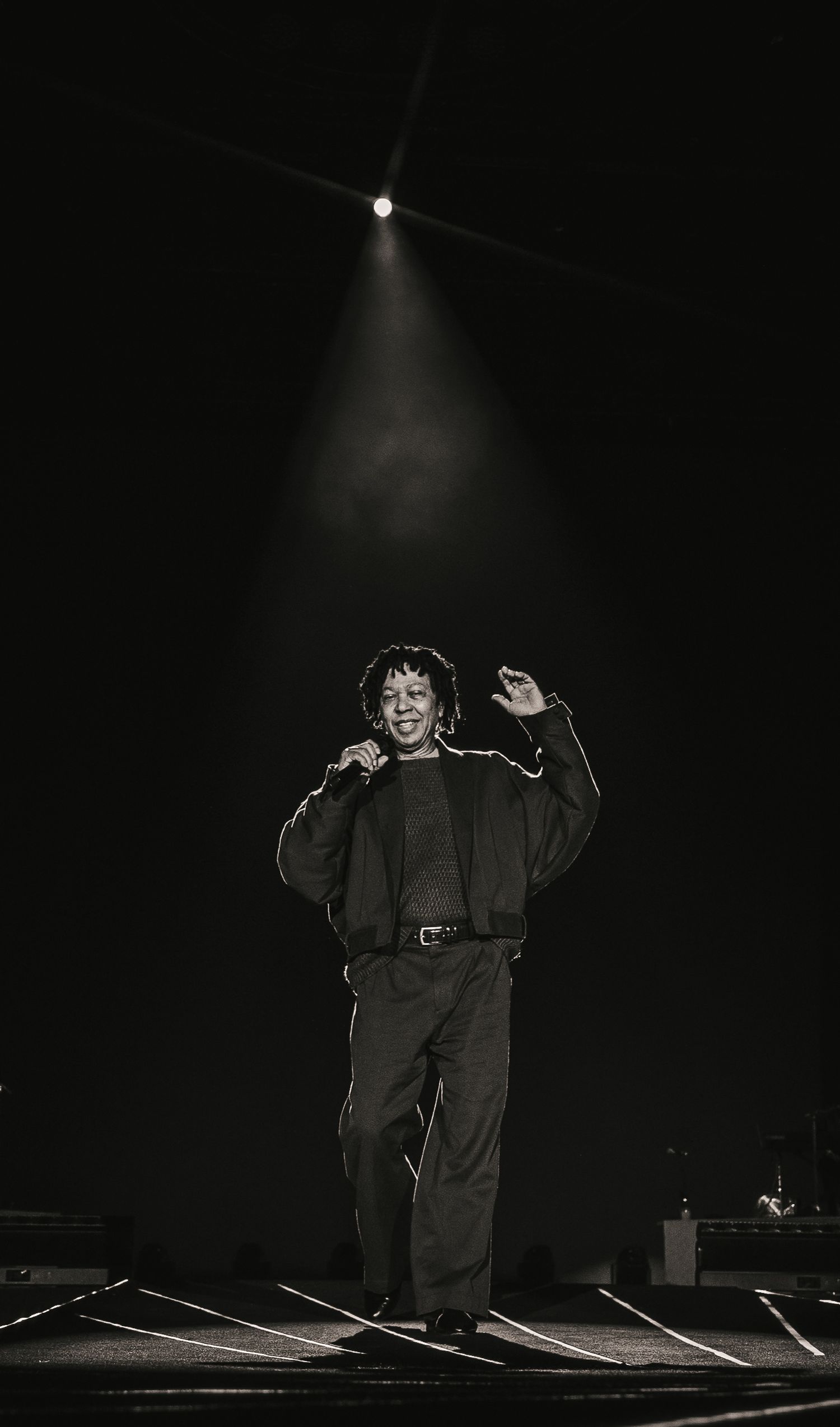 Black and white photo of a performer singing on stage under a dramatic spotlight beam in a dark concert venue.