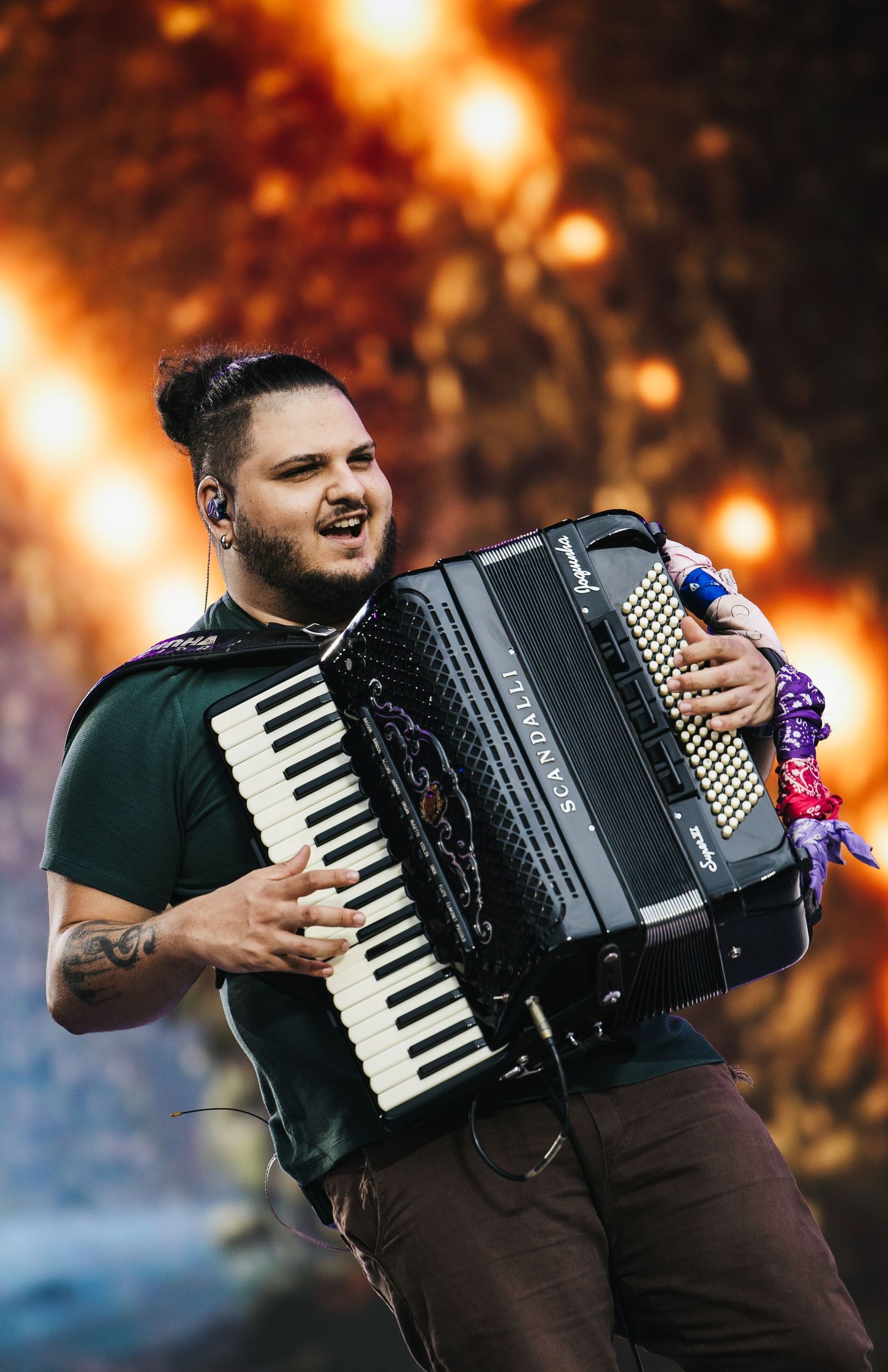 Musician playing a black piano accordion on stage with dramatic orange lighting behind him.