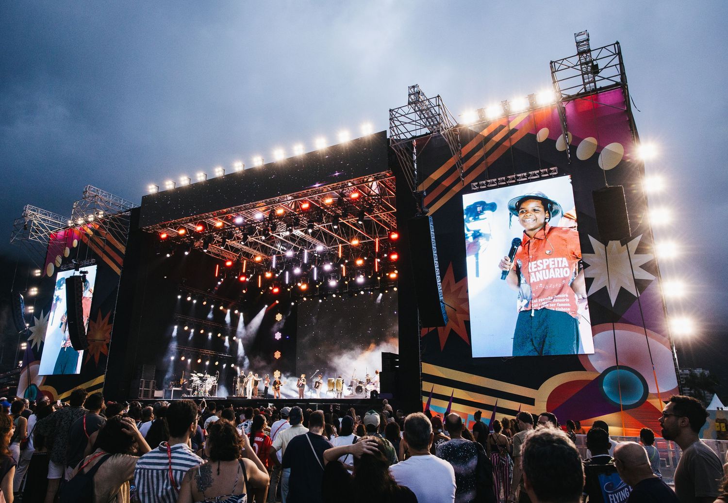 Large outdoor music festival stage with bright lights, big screens, and a crowd enjoying a live concert performance.