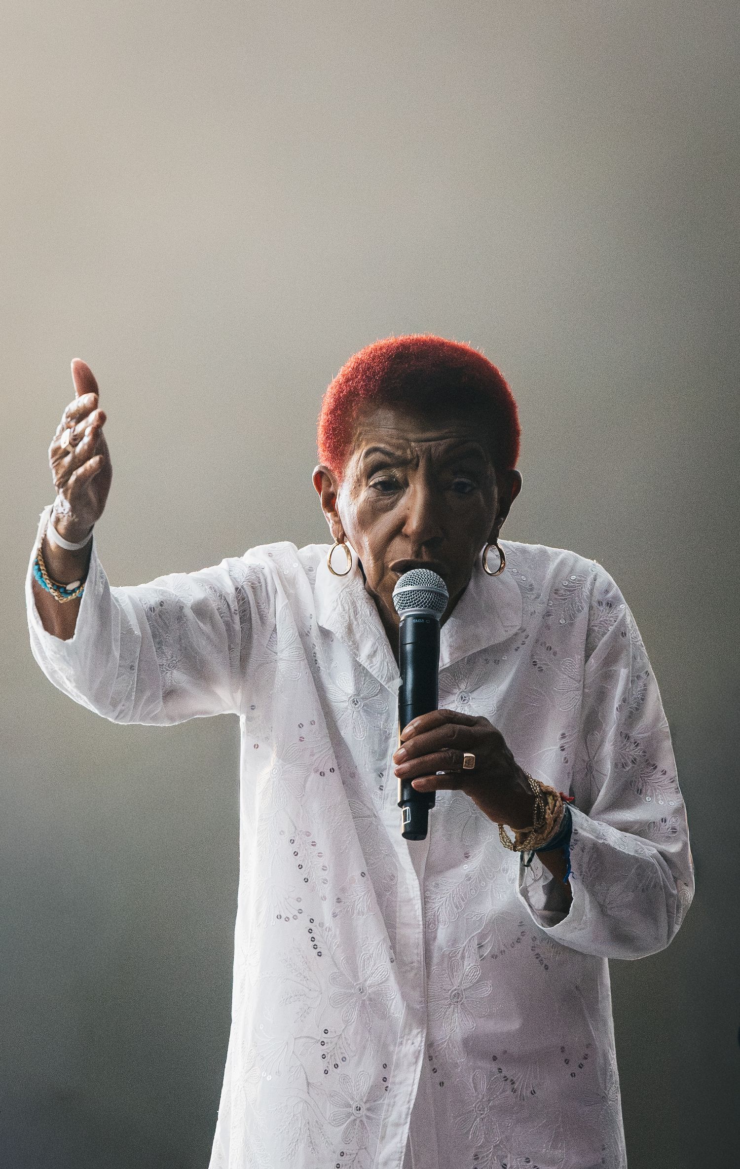 Elderly woman with red hair singing into microphone, wearing white jacket, raising hand on stage.