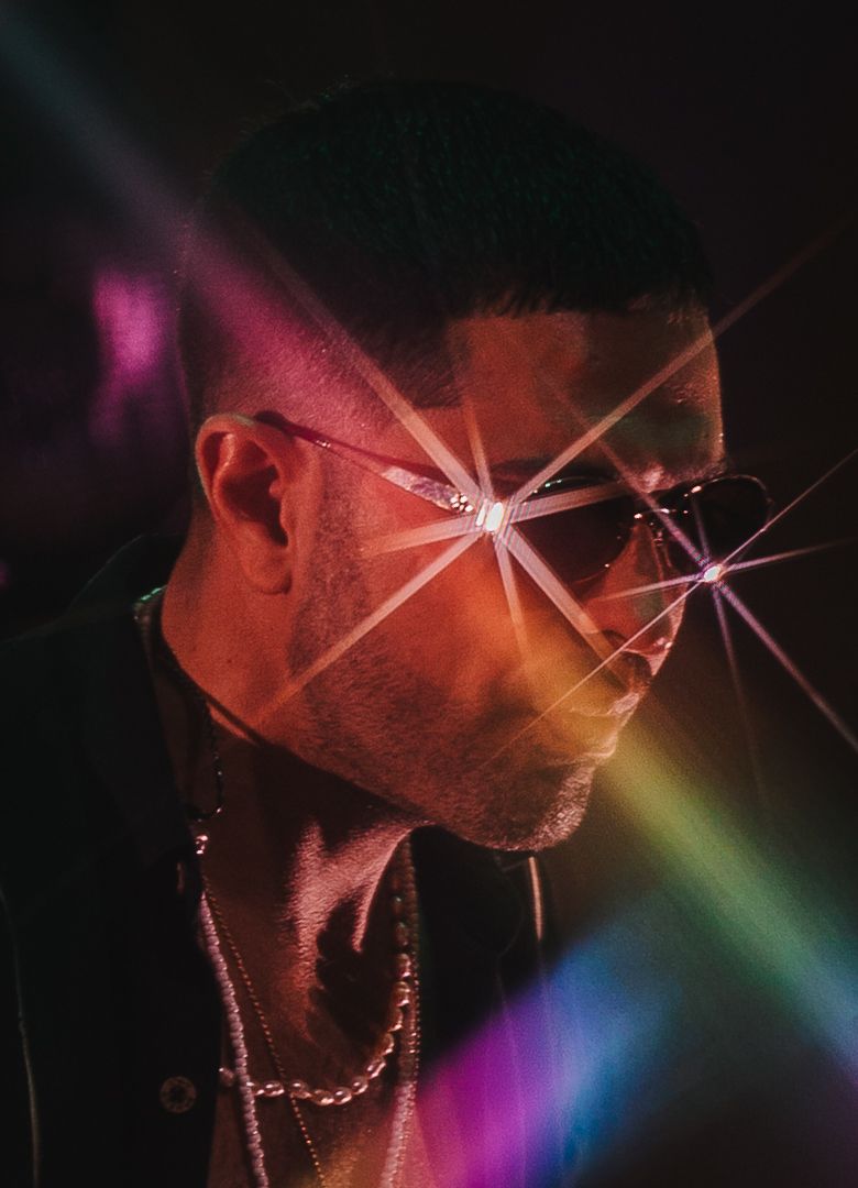 Male artist wearing sunglasses with rainbow lens flare effect under colorful concert stage lighting.