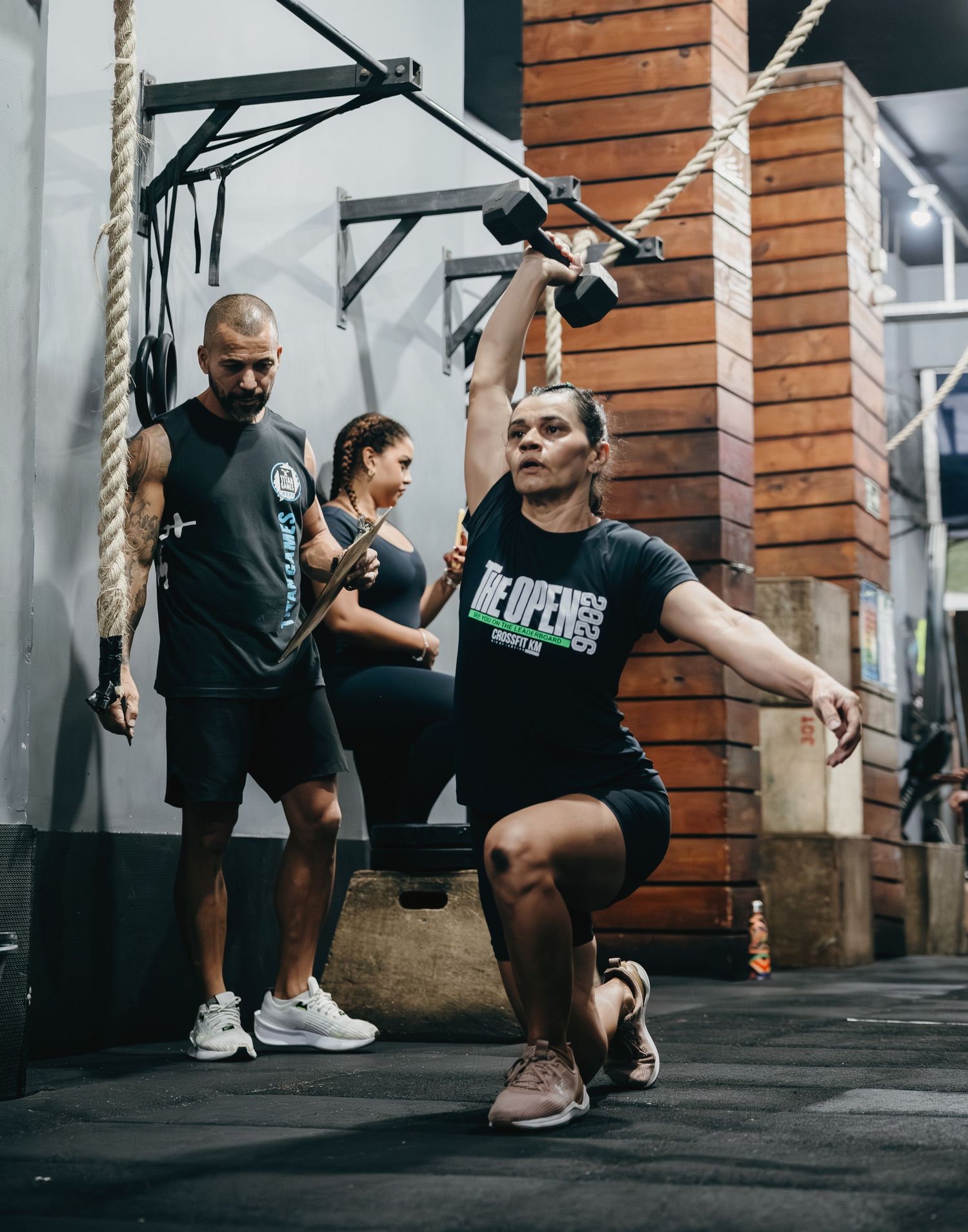 Woman performs overhead dumbbell squat at CrossFit gym while coach observes during The Open competition.