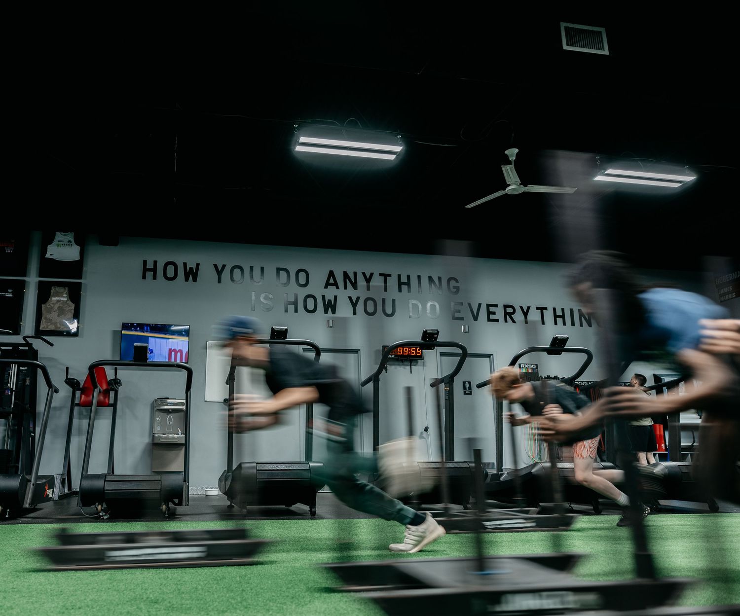People running on treadmills in a dark gym with motivational quote on wall reading How You Do Anything Is How You Do Everything.