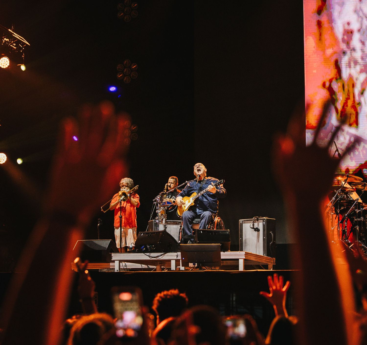 Live concert performance with musicians on stage playing guitar while excited crowd raises hands in a dark arena.