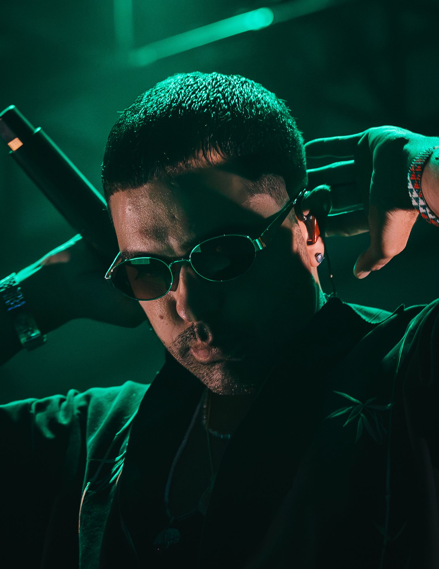 Male performer in sunglasses poses dramatically under green stage lighting at a live concert.