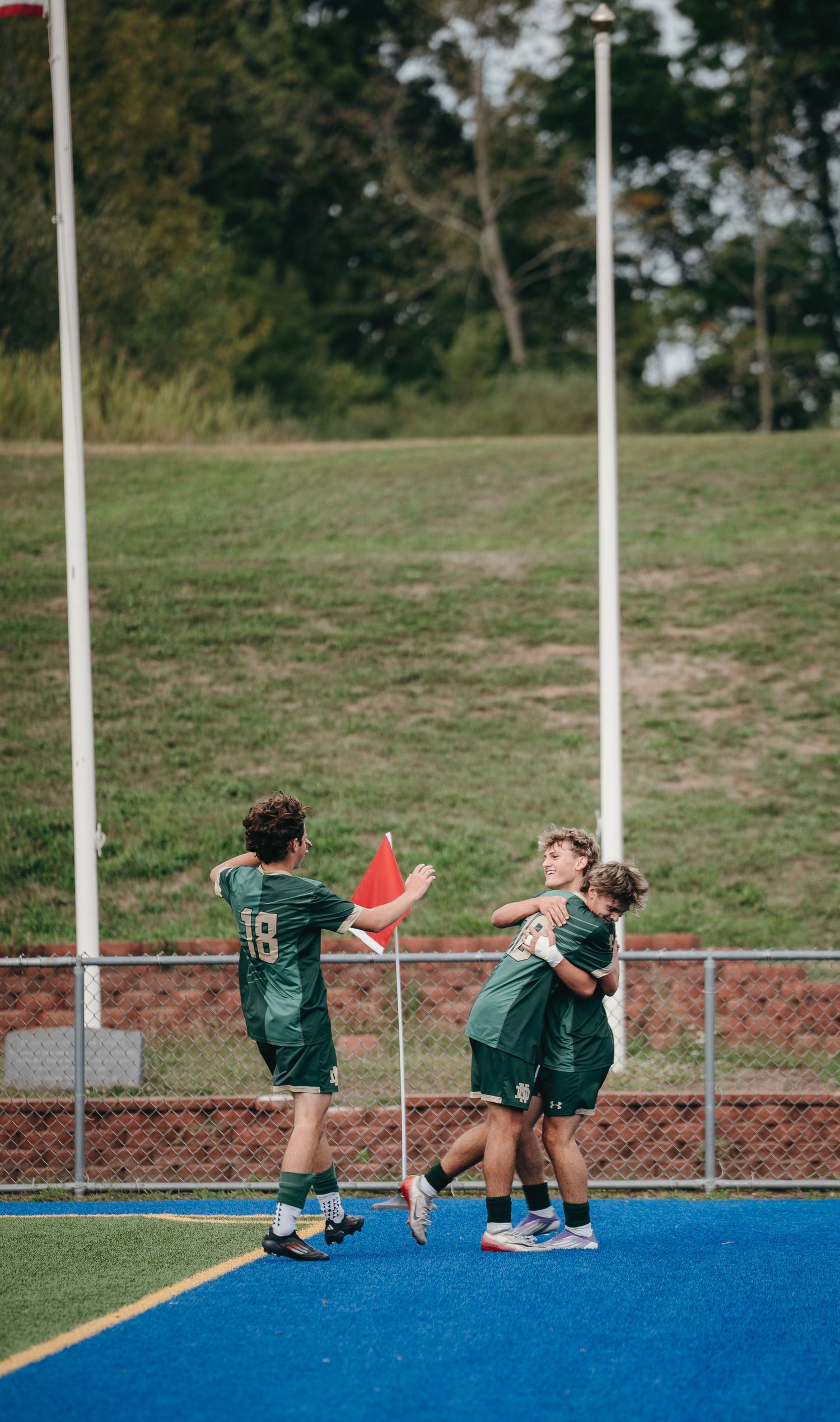 Three youth soccer players in green uniforms celebrate on a blue turf field near a corner flag.