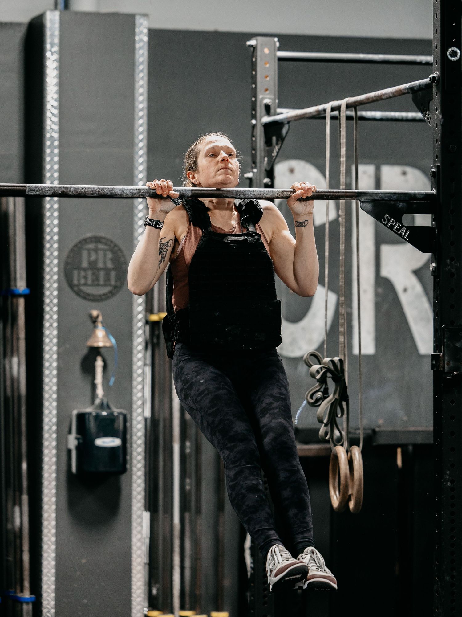 A muscular woman performs pull-ups on a bar in a CrossFit gym, wearing a black tank top and camo leggings.