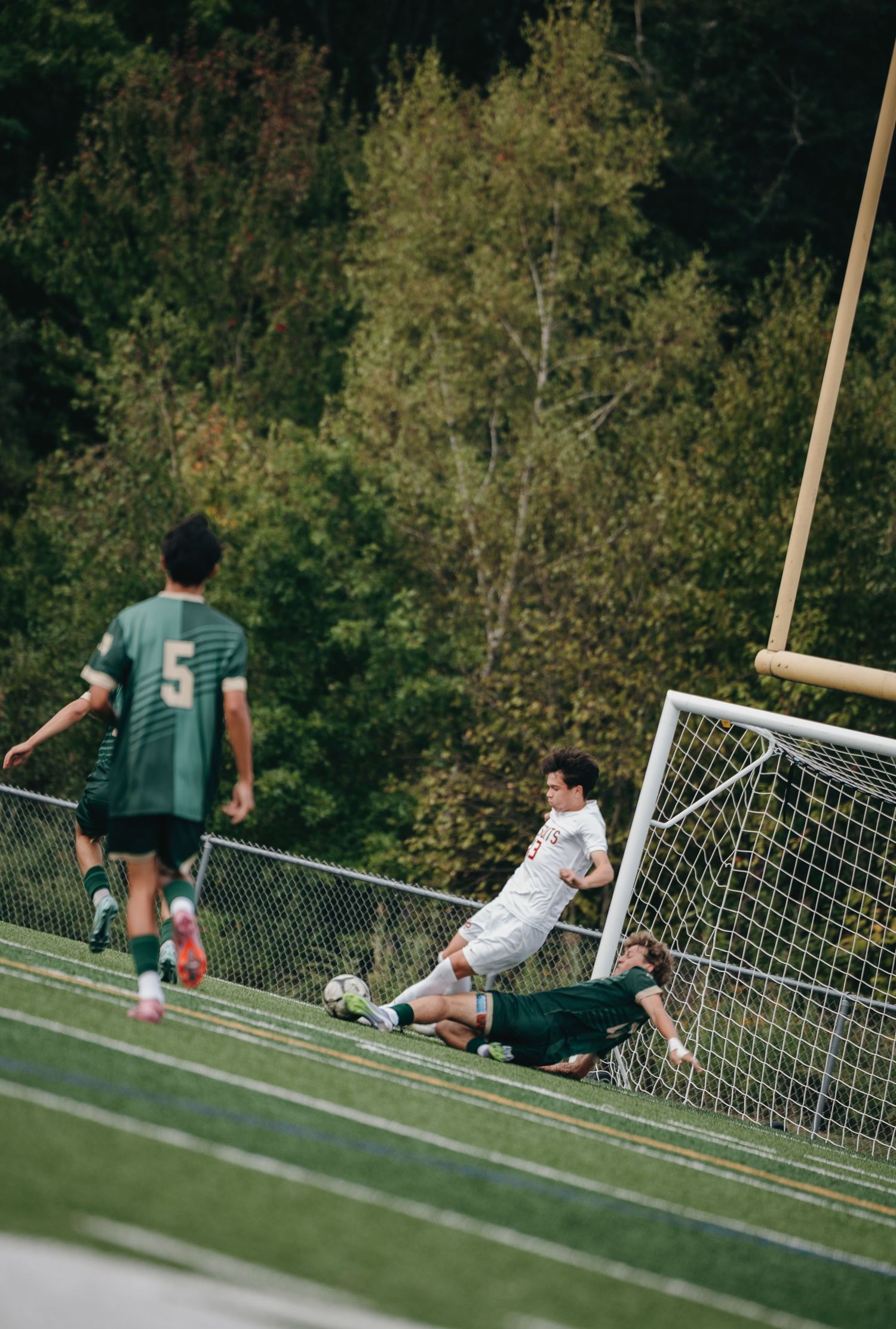 Soccer players compete near the goal on a green turf field, with one player sliding and another in white defending.