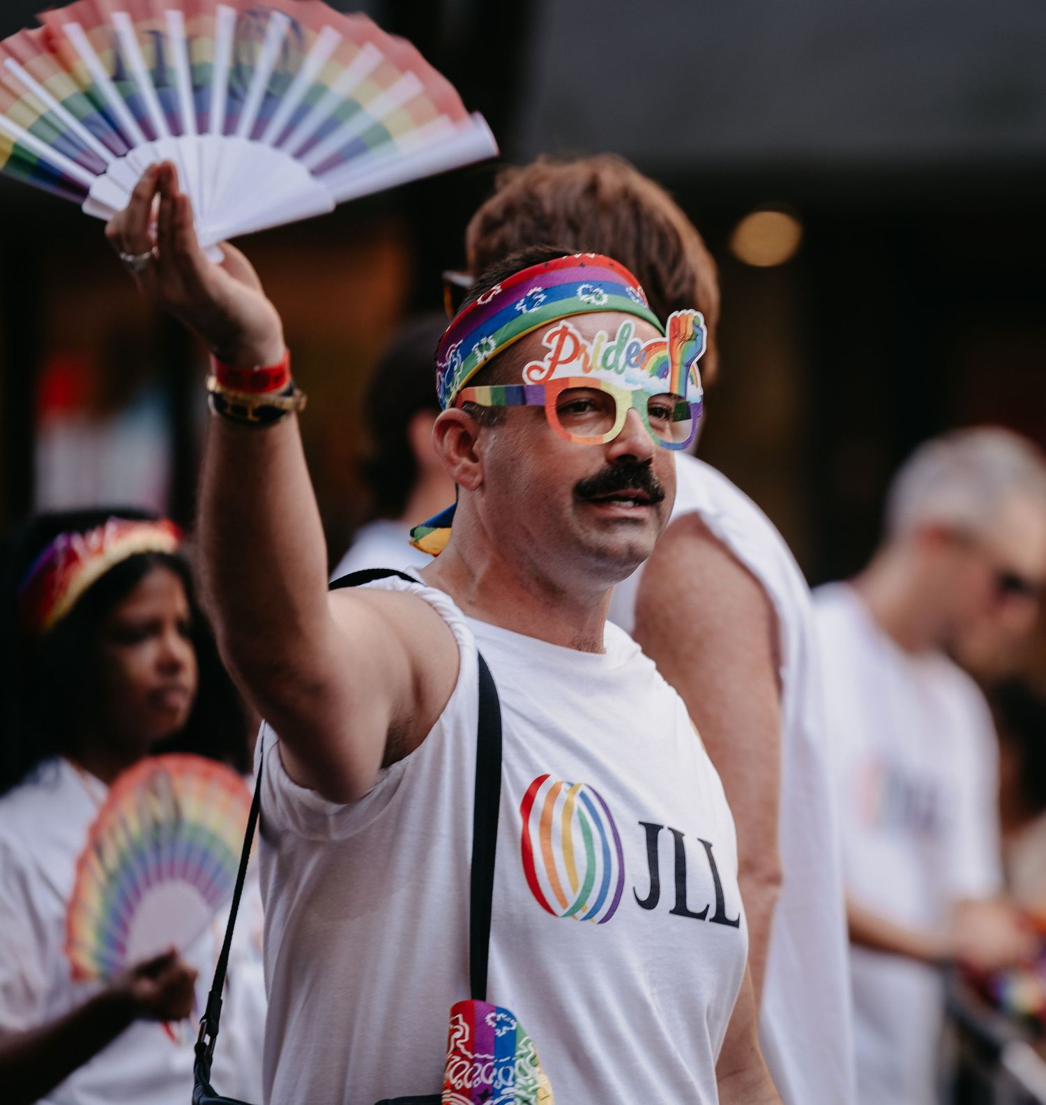 Man in Pride parade wearing rainbow accessories and JLL shirt holding colorful fan.