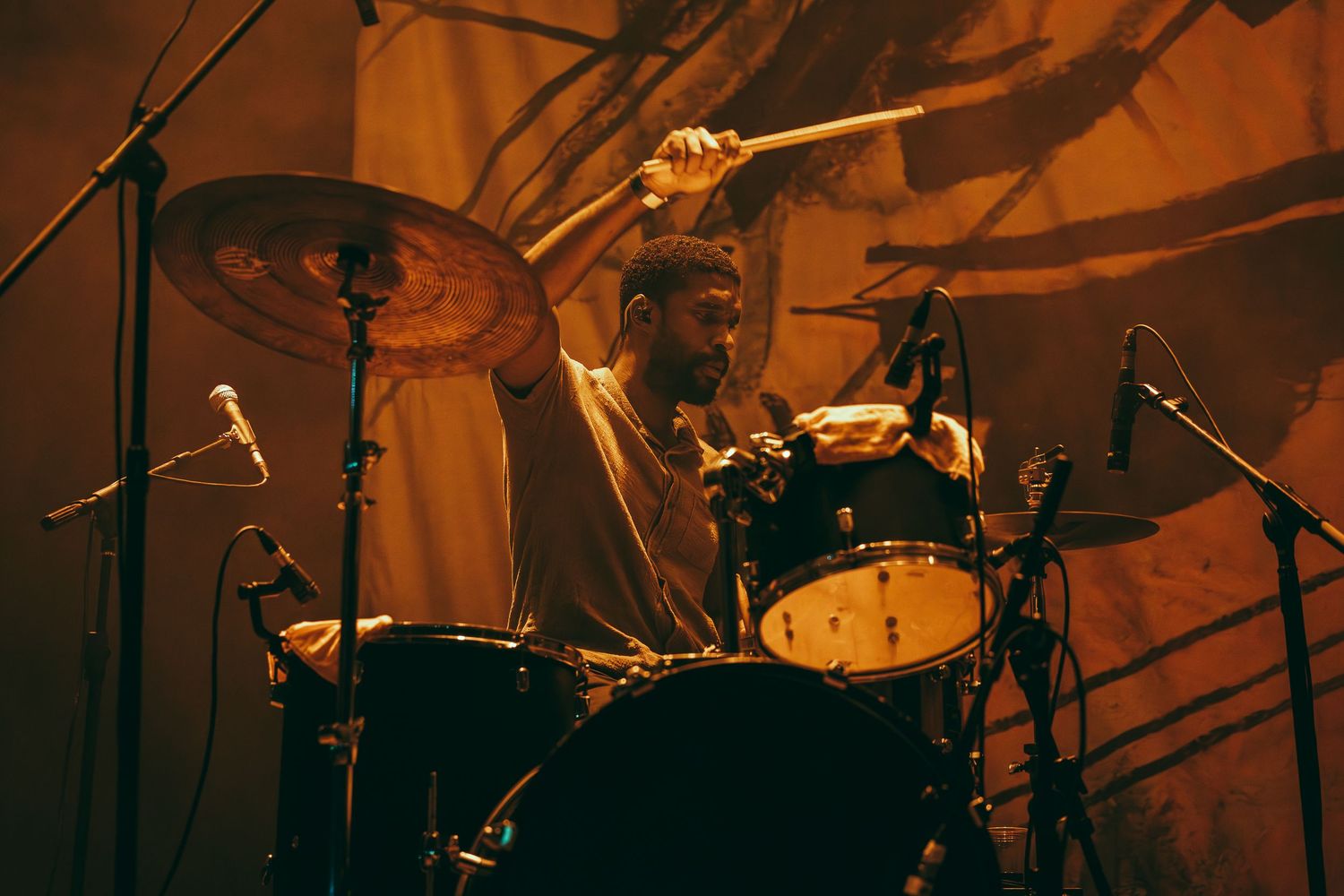 A drummer performs energetically on stage under warm amber lighting at a live music concert.