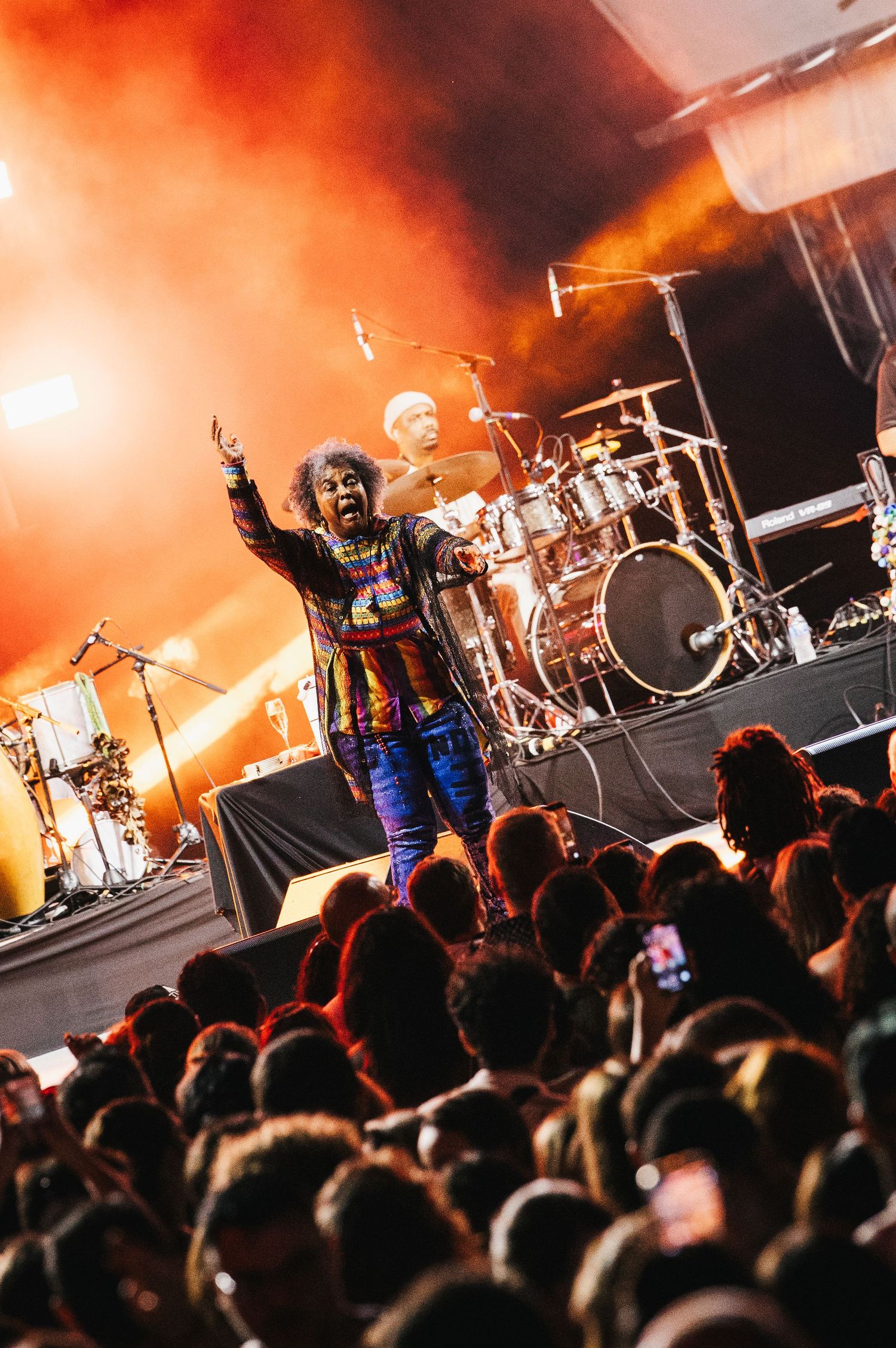 Energetic performer in colorful outfit raises hand to crowd at vibrant outdoor concert with dramatic orange stage lighting.