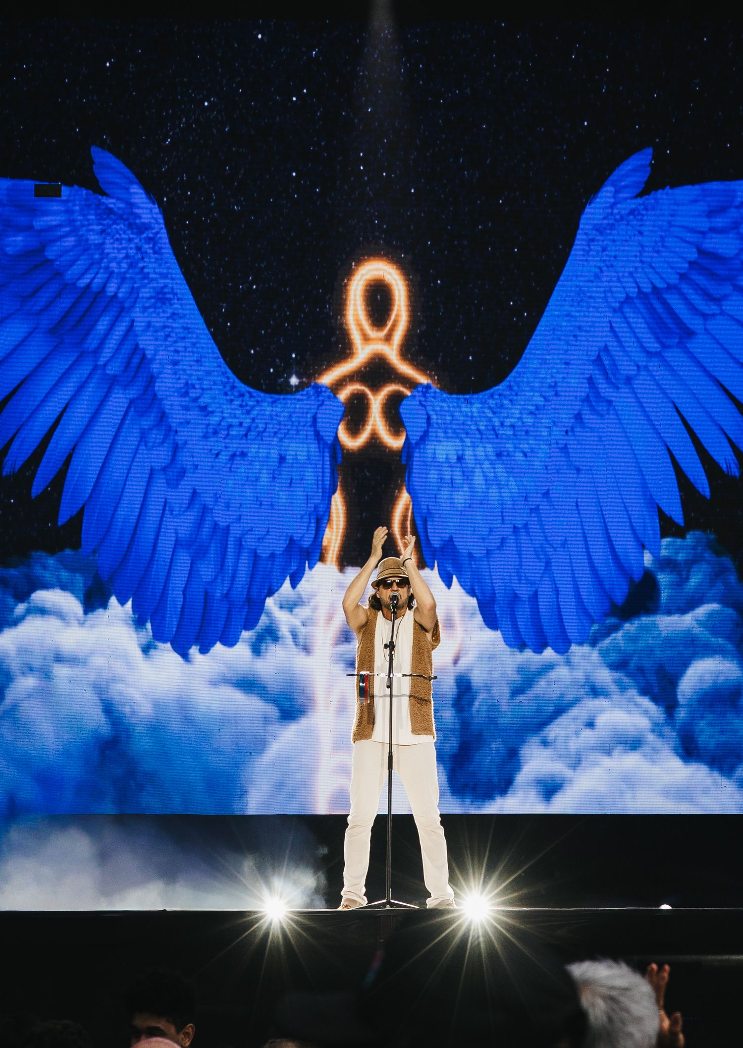 Performer stands at microphone on stage with giant blue angel wings and glowing symbol backdrop.
