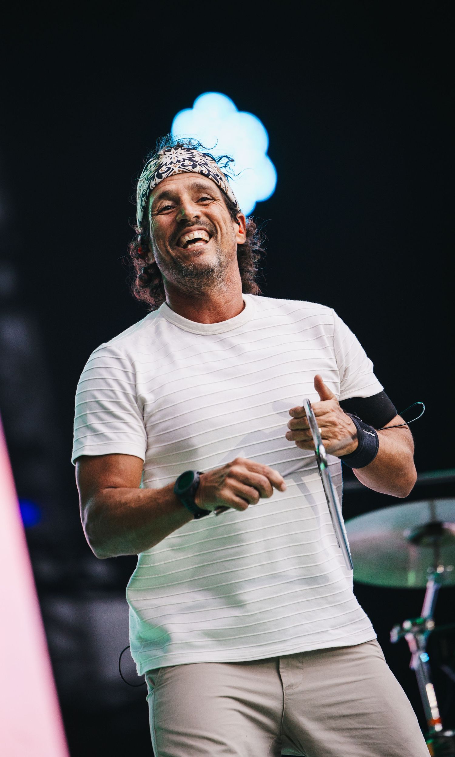Smiling male performer on stage wearing a white t-shirt and bandana headband, holding a microphone at an outdoor concert.