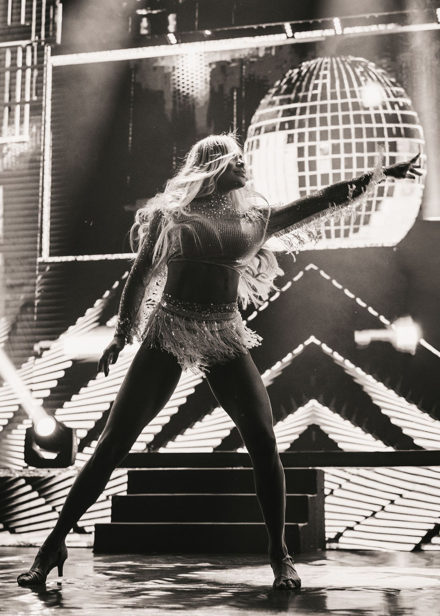 A glamorous dancer poses dramatically on stage with a disco ball backdrop in a black and white photo.