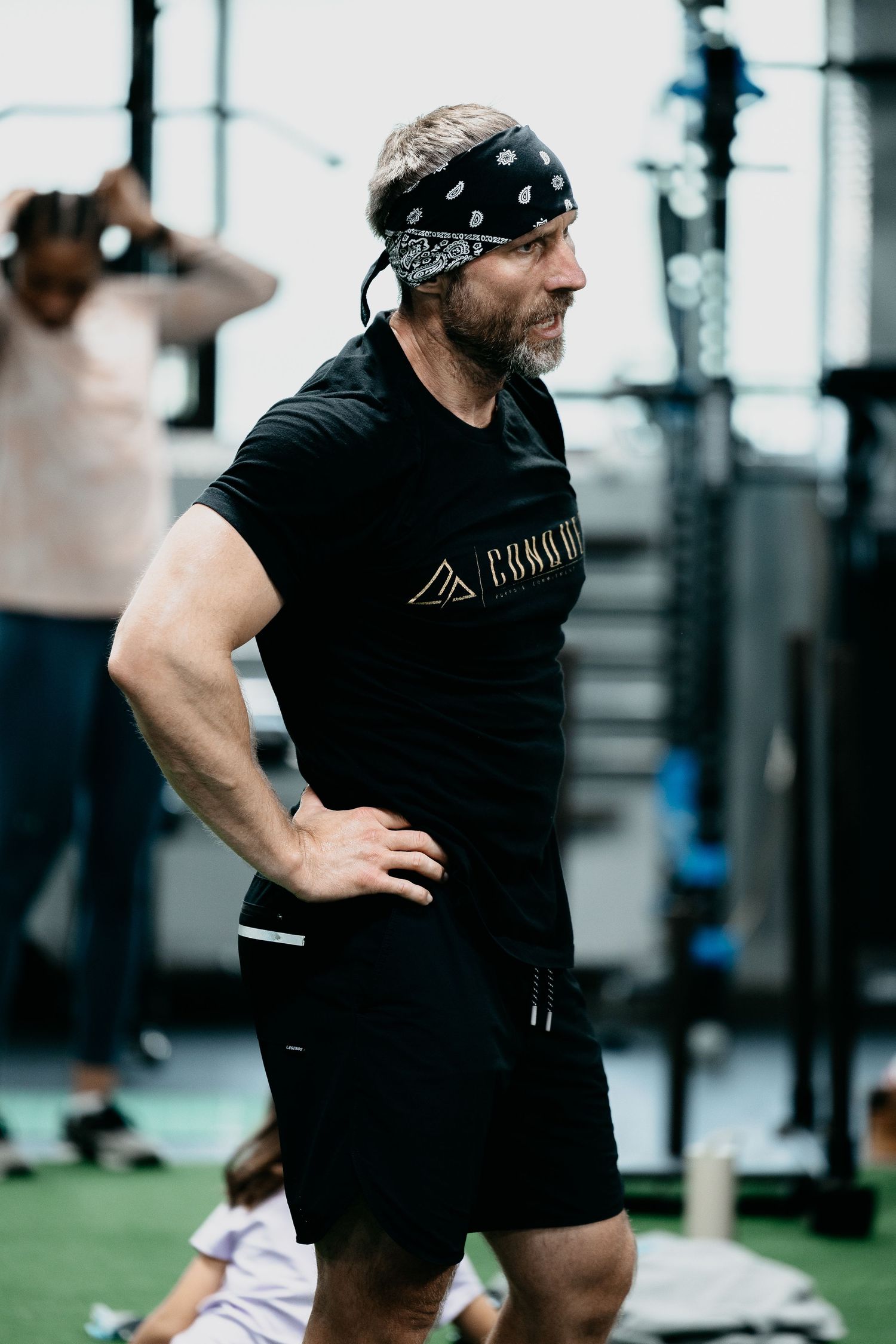 Muscular man in black t-shirt and bandana headband stands confidently in a gym setting.