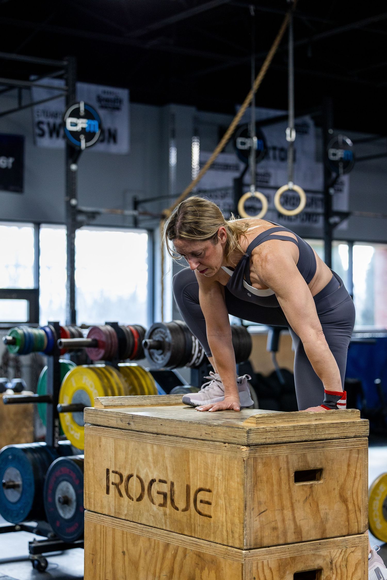 Woman performing box jump exercise on Rogue wooden plyo box in CrossFit gym with barbells and rings.