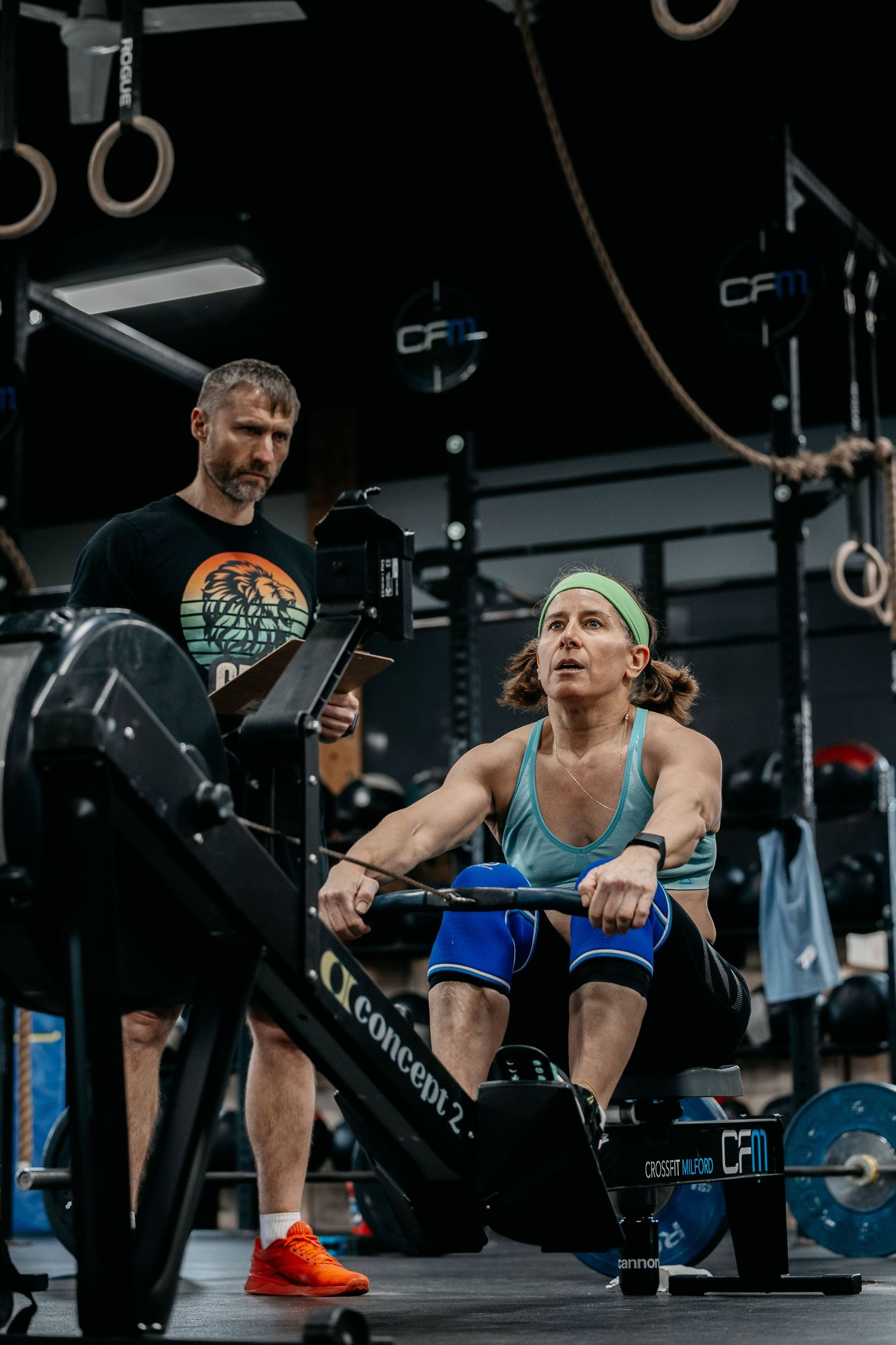 Woman using Concept2 rowing machine in CrossFit gym while male coach observes during intense workout session.