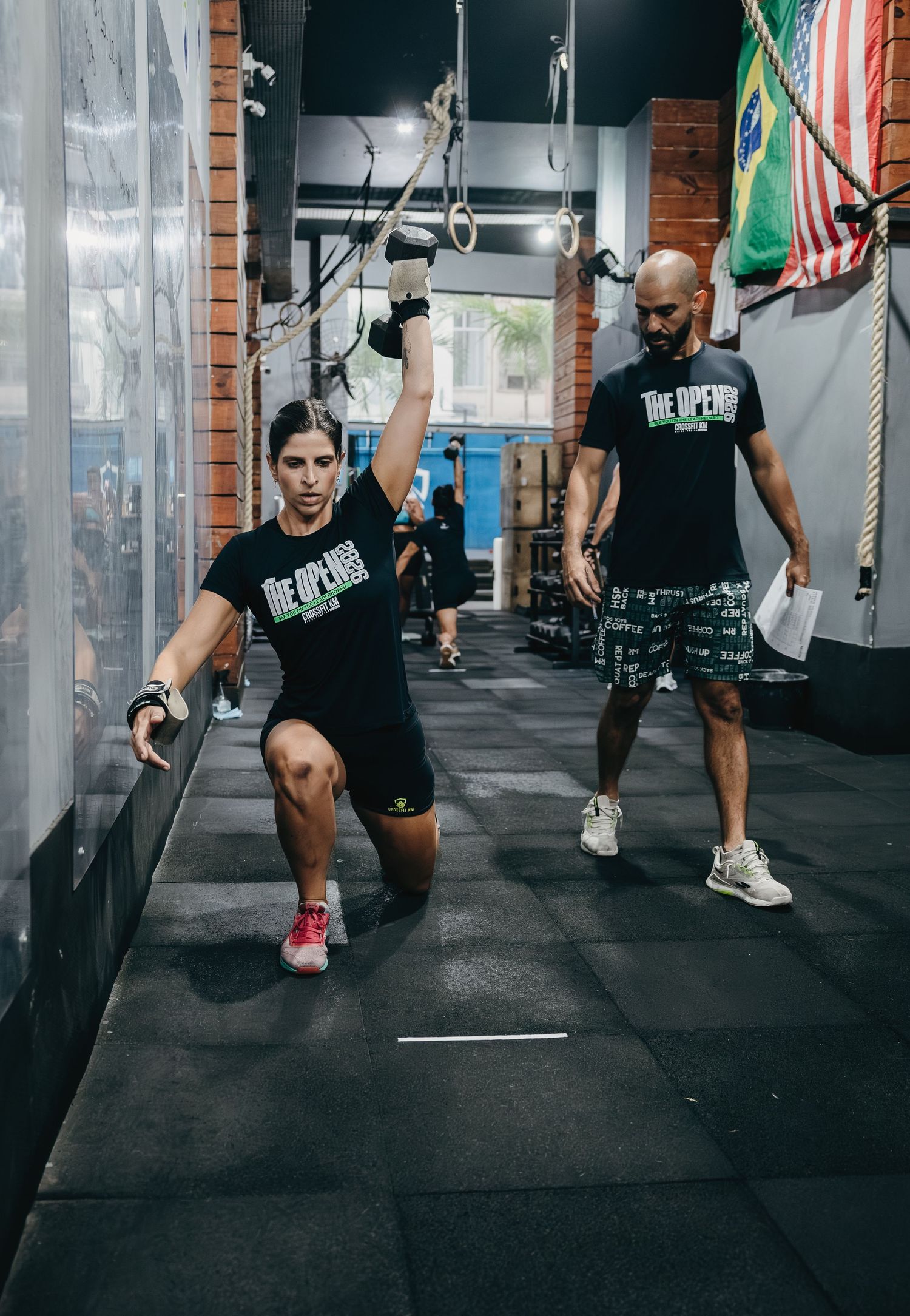 Woman doing a lunge exercise in a CrossFit gym while a trainer observes during The Open competition.