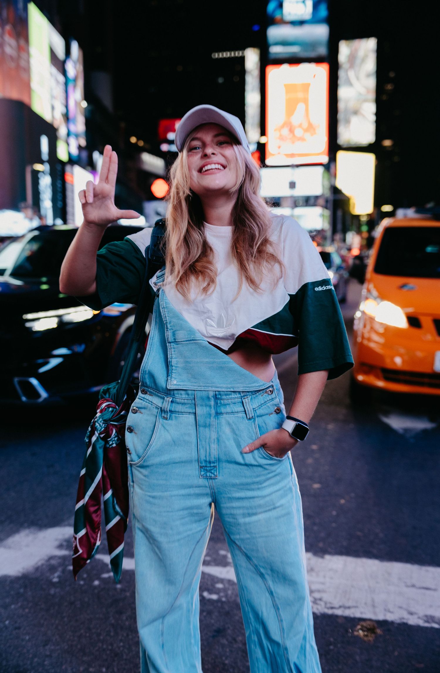 Smiling woman in overalls and white cap posing at Times Square NYC at night.