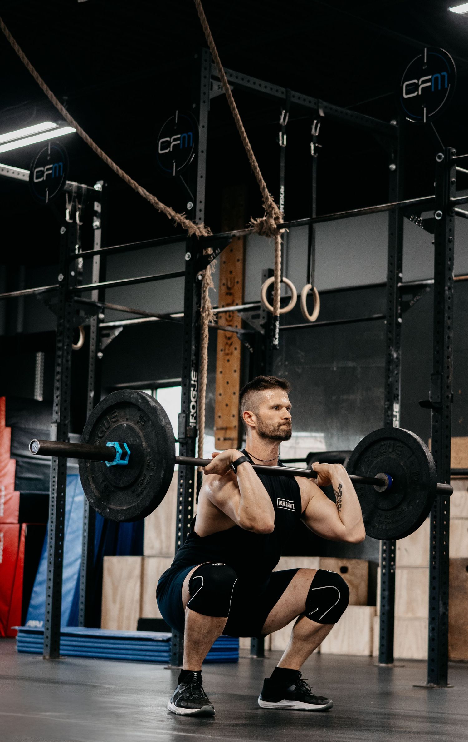 Muscular man performing a barbell back squat in a CrossFit gym with ropes and gymnastic rings.