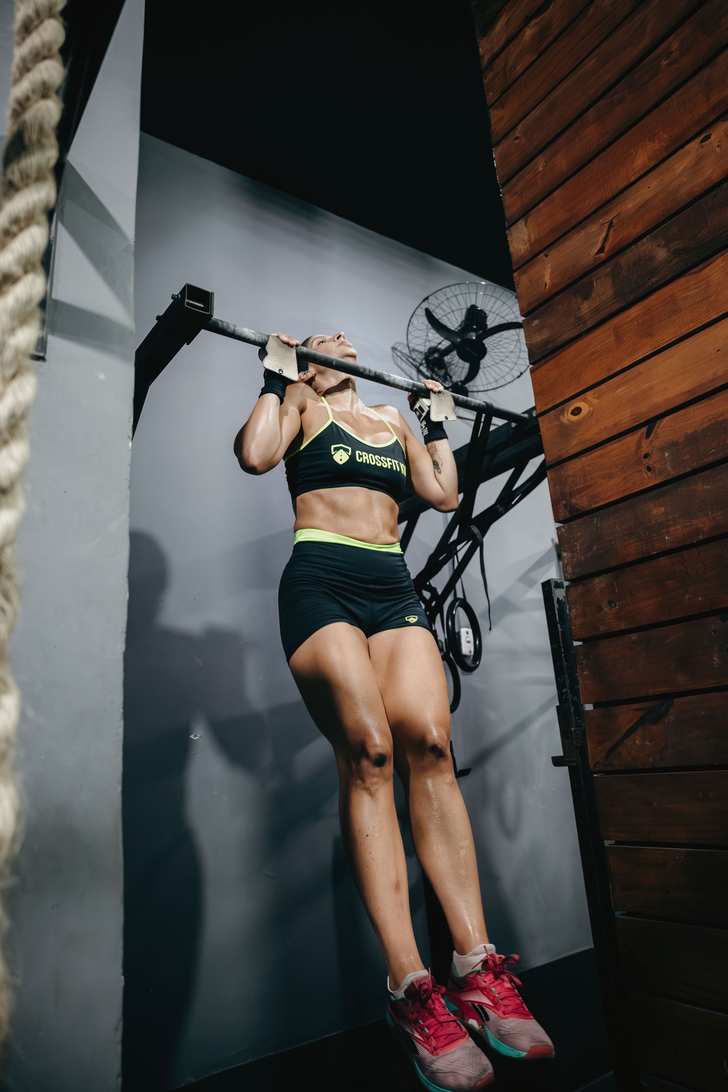 Woman in CrossFit gear performing pull-ups on a bar in a gym with wooden walls and a fan.